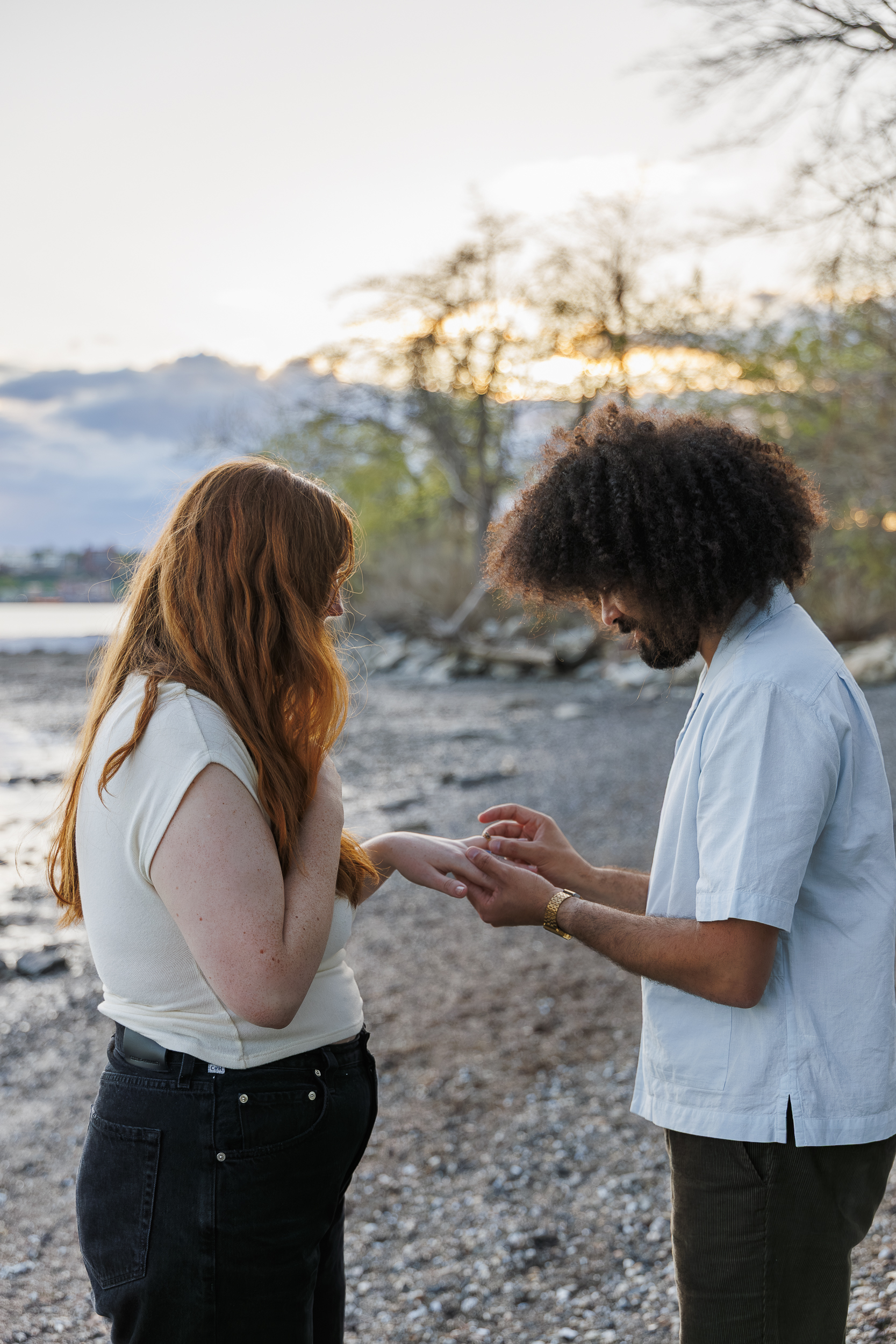 A man places a ring on a woman's finger while standing together on a rocky beach at sunset, capturing their plan proposal in stunning photography with trees and water in the background.