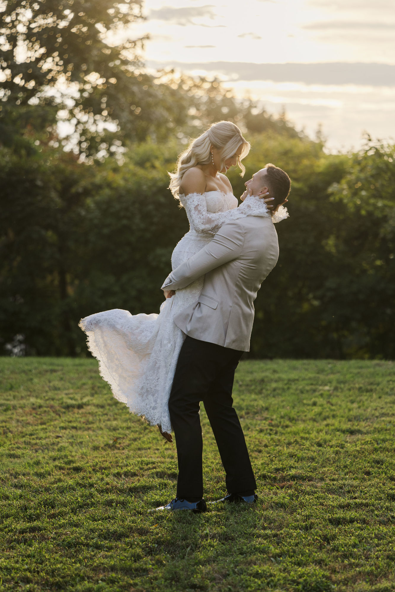 A groom in a light jacket lifts his bride, dressed in a white lace gown, as they smile at each other outdoors on a grassy field—capturing pure joy as everything on their wedding day checklist falls perfectly into place at sunset.