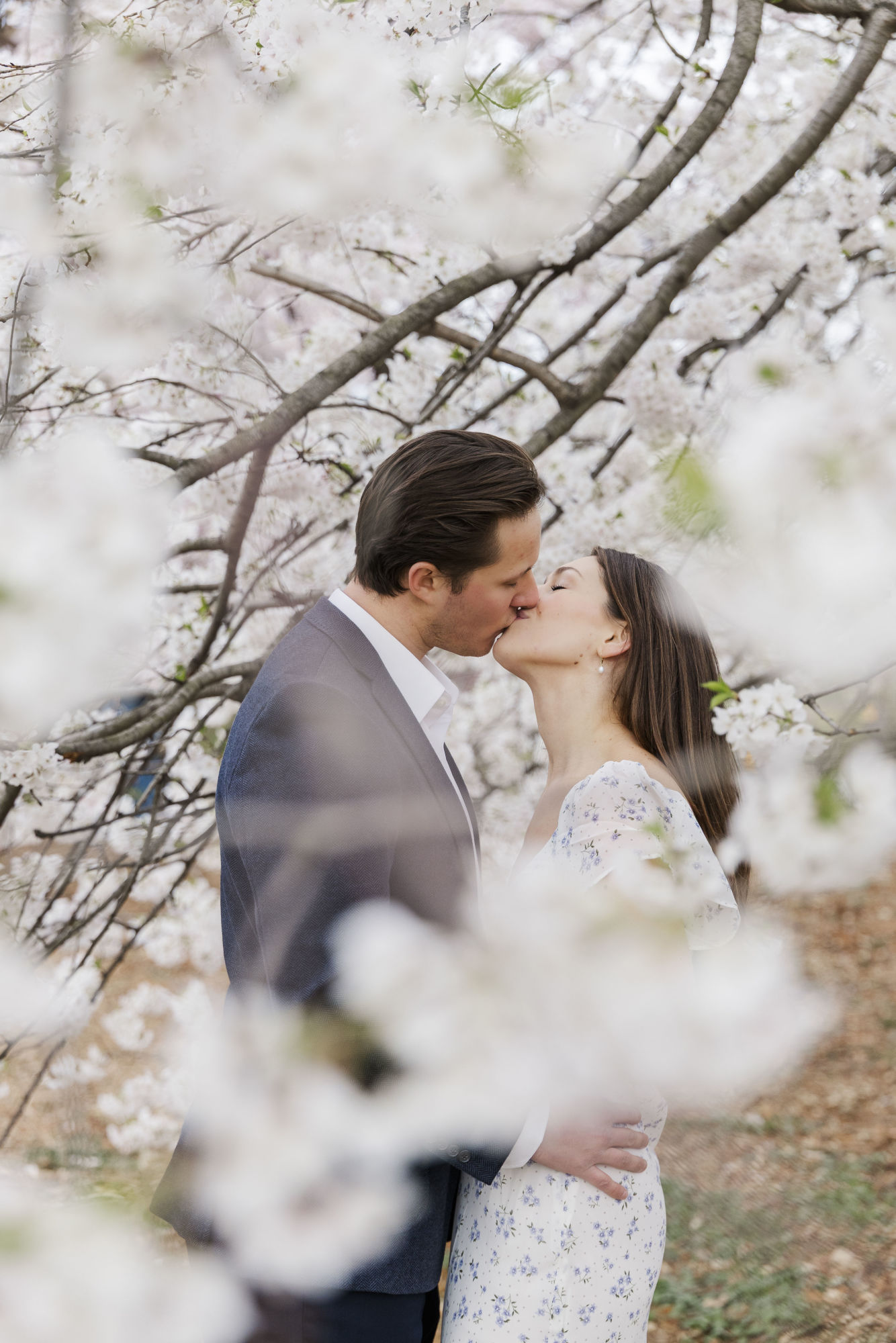 A couple kisses tenderly under blossoming cherry trees, surrounded by soft white flowers in Central Park. The man wears a suit and the woman a light, floral dress—capturing romantic and serene Central Park engagement photos.