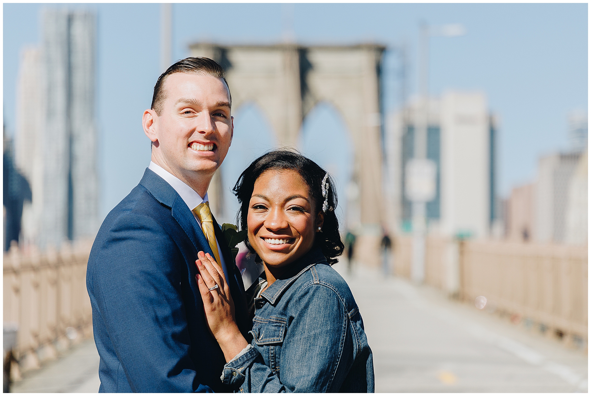 A smiling couple poses together on the Brooklyn Bridge, dressed for their wagner cove elopement—the man in a blue suit and the woman in a denim jacket. The city skyline glows in the background on a sunny day.