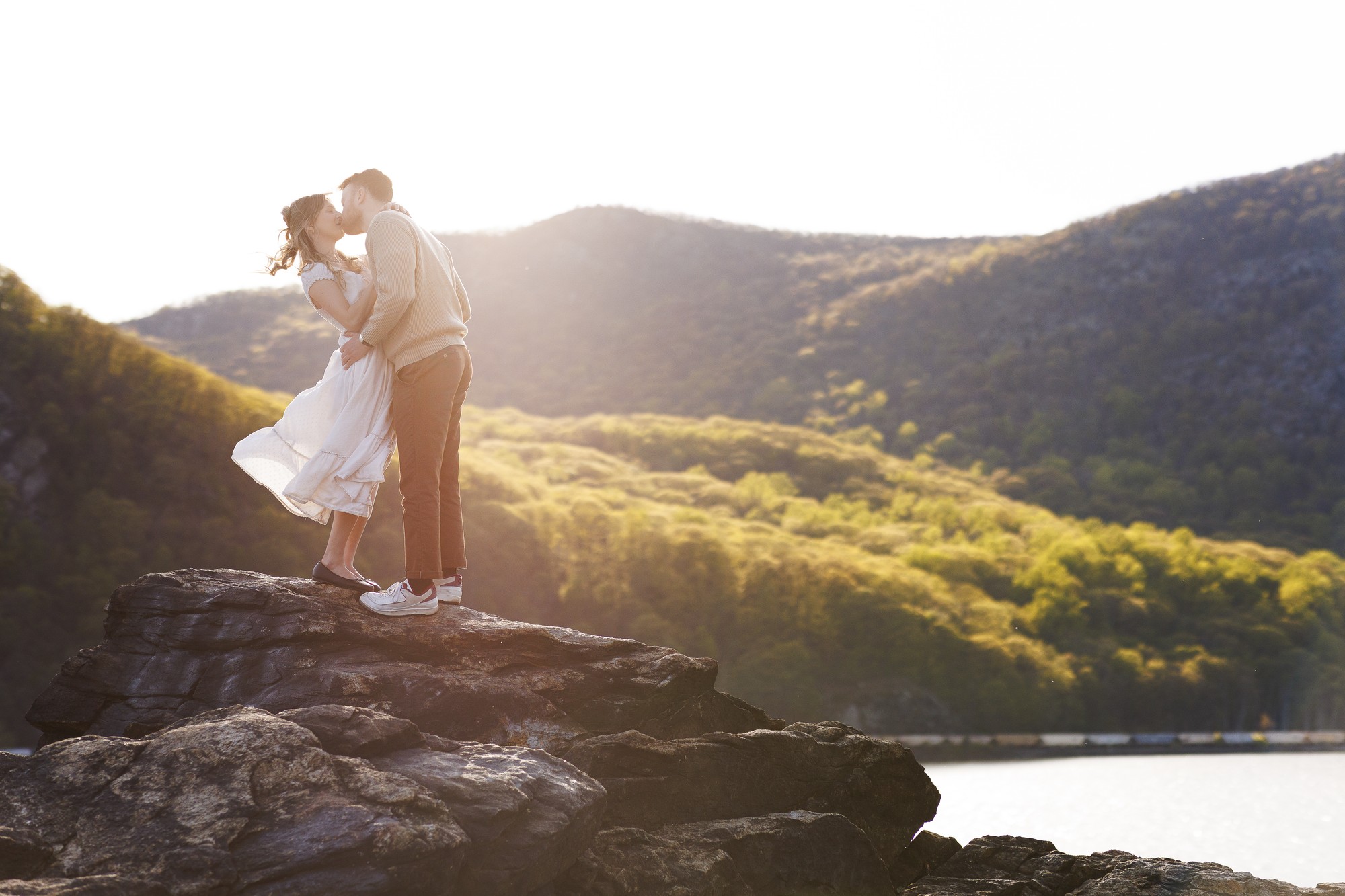 Couple kissing on rocky cliff overlooking Hudson Valley landscape, Hudson Valley wedding photography moment captured during golden hour sunset