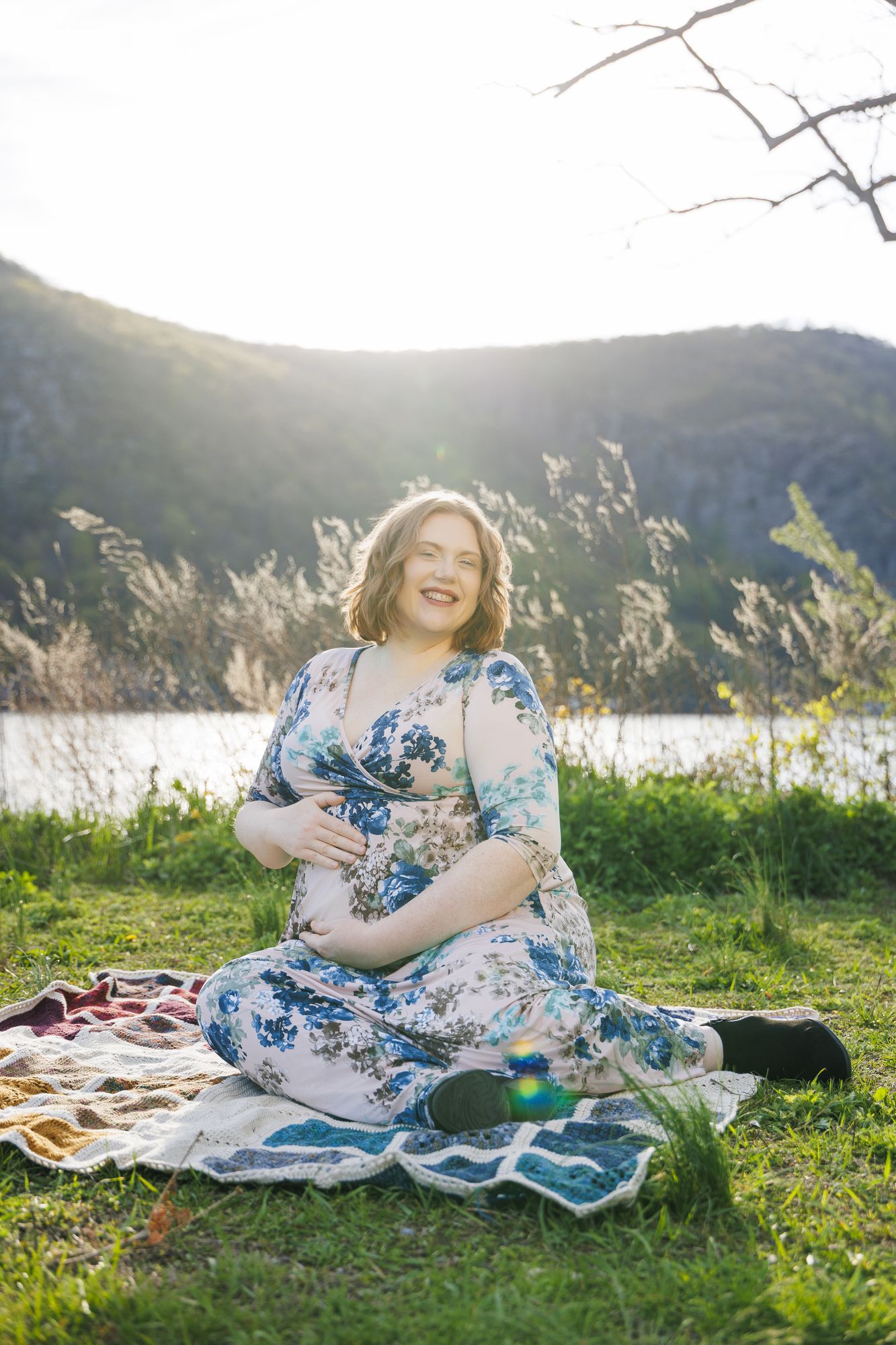 Pregnant woman in floral dress sitting by lake with mountains, captured by Hudson Valley wedding photography team specializing in maternity and portrait sessions.