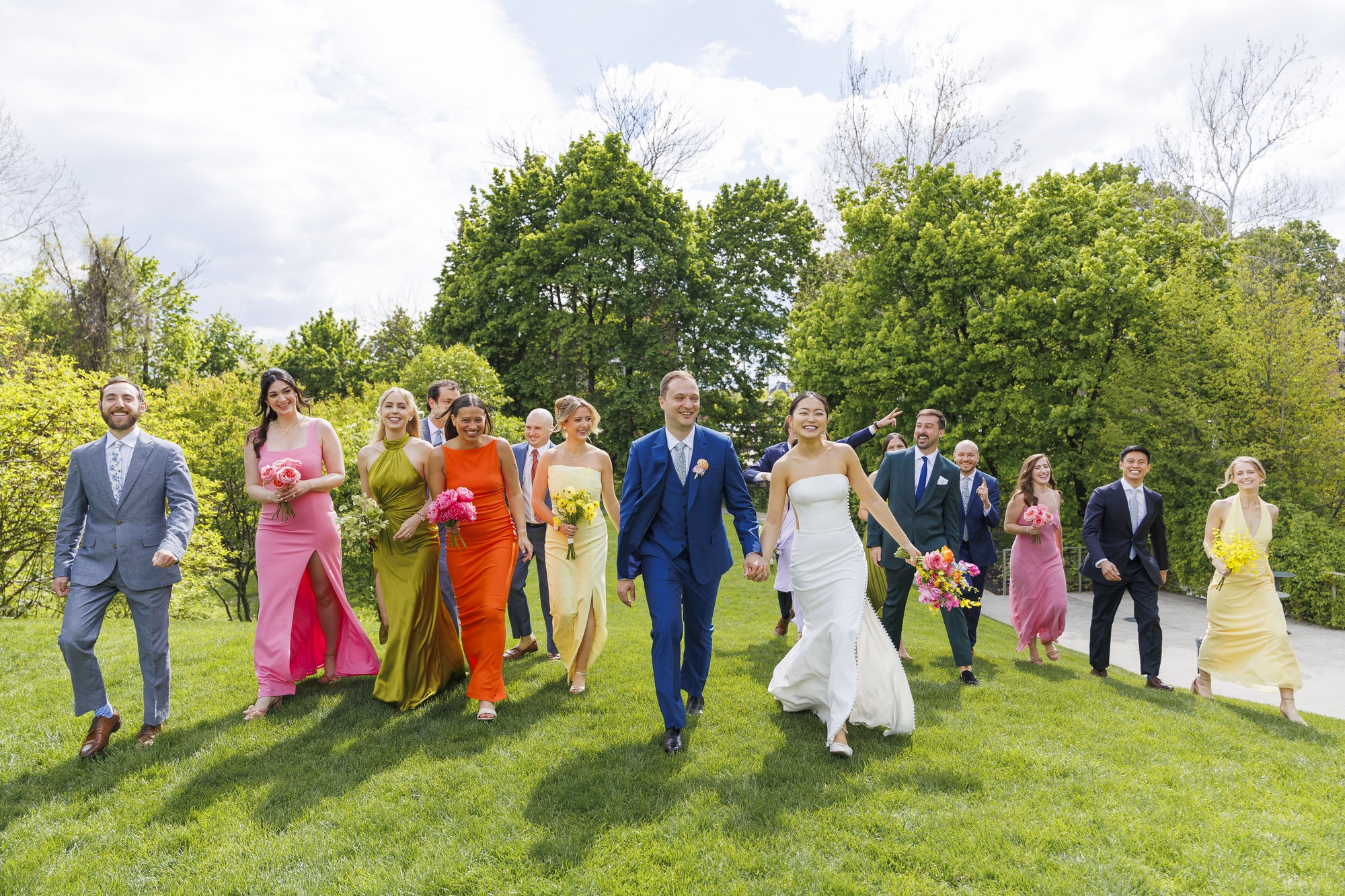 A bride and groom walk hand in hand outside with their wedding party, all dressed in colorful formalwear, smiling as they stroll across a green lawn at their roundhouse wedding beneath a partly cloudy sky.