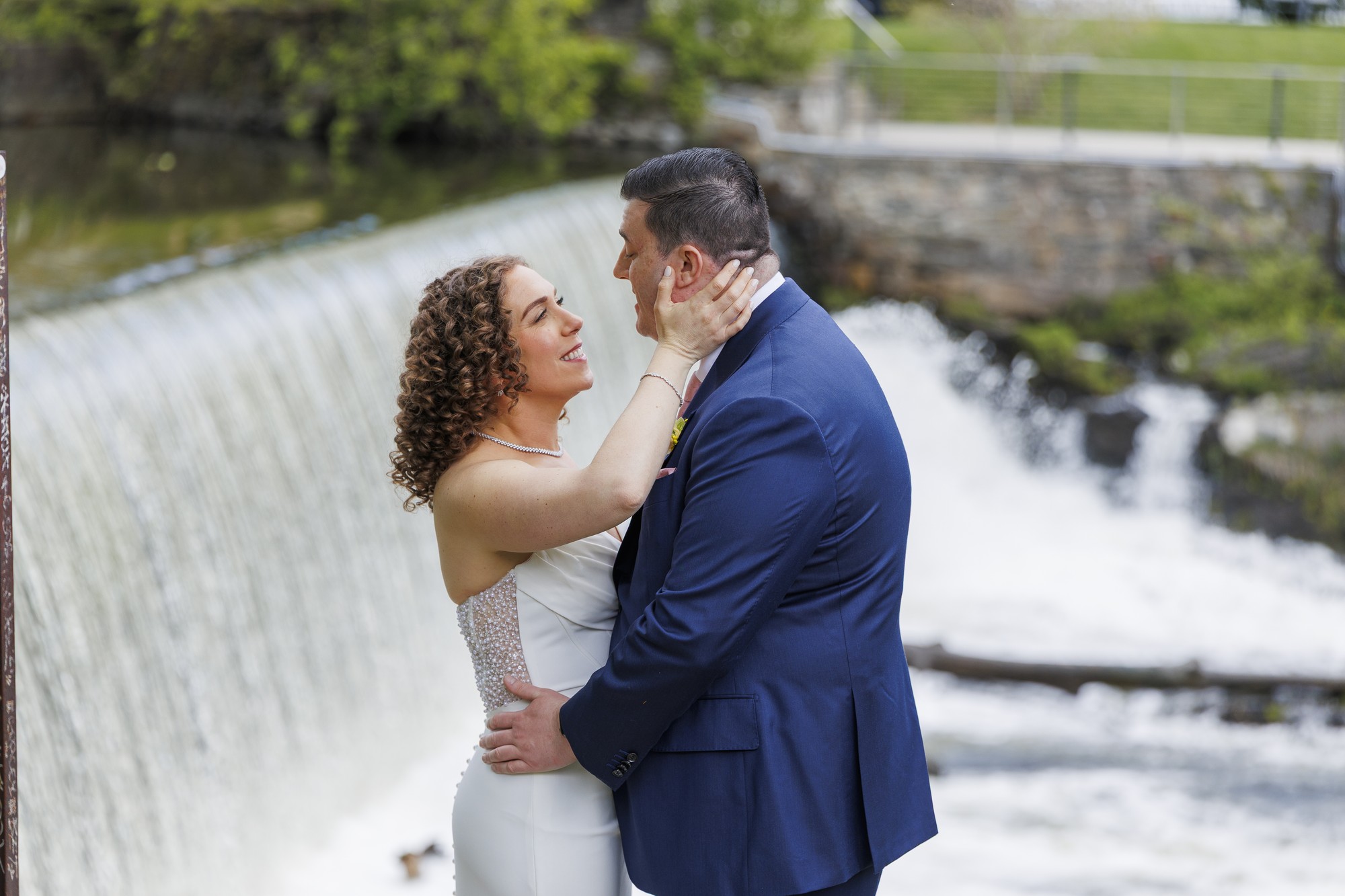 A bride and groom embrace and smile at each other in front of a cascading waterfall, surrounded by greenery during their wedding at the Roundhouse. The bride wears a white dress and the groom is in a navy suit.