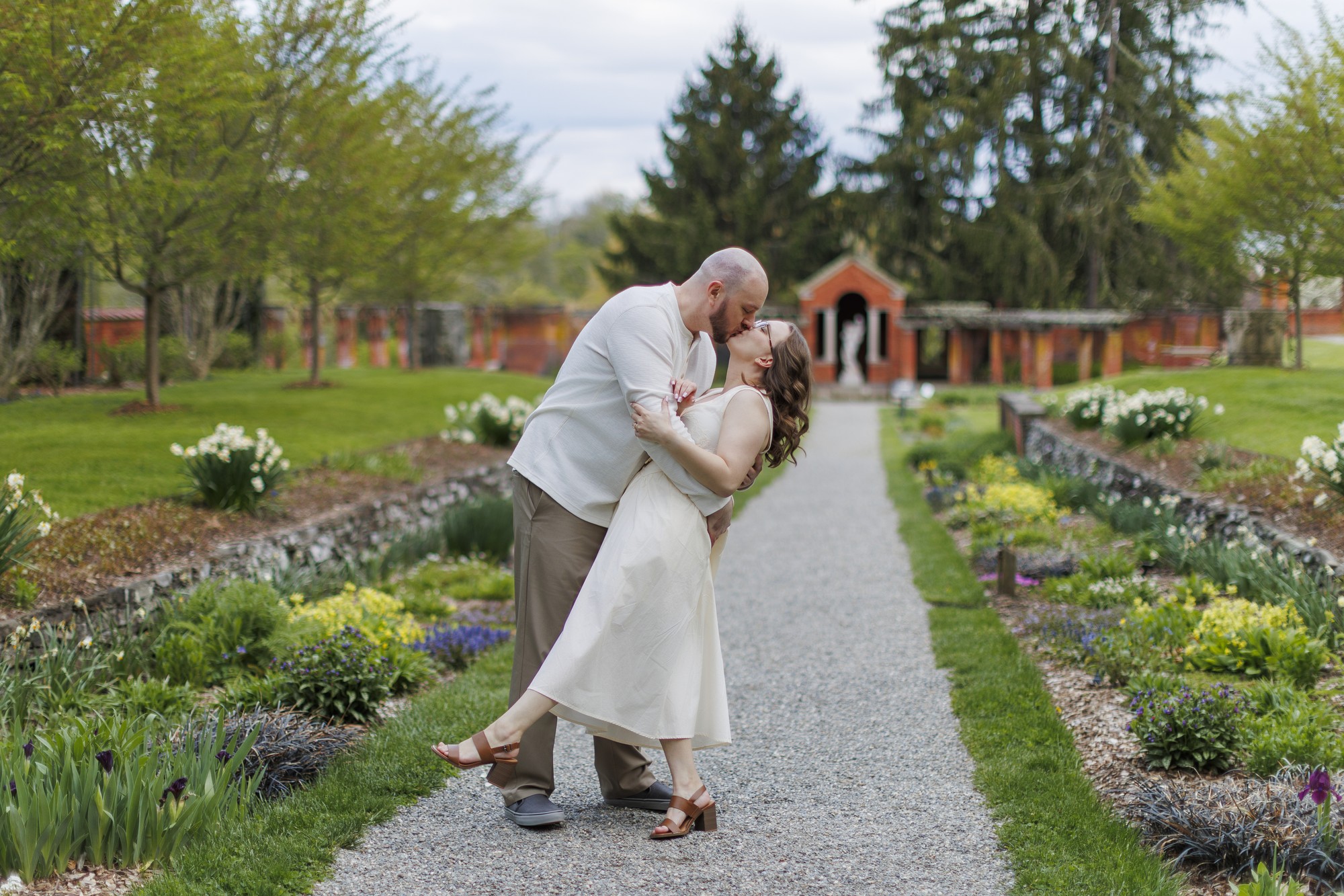 A couple stands on a garden path at Vanderbilt Mansion; the man dips the woman as they smile and embrace, surrounded by lush greenery, blooming flowers, and trees under a cloudy evening sky.