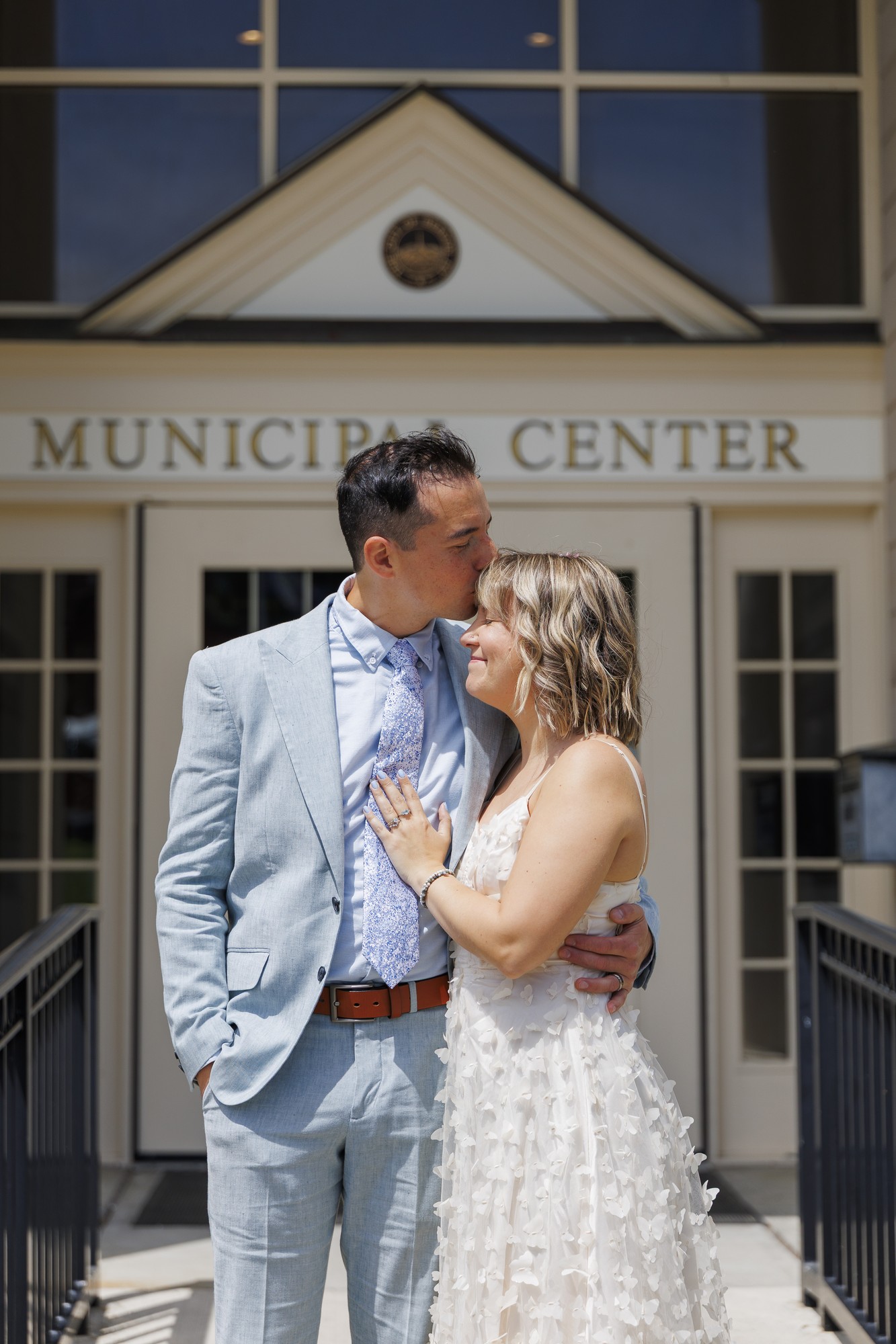 A couple stands embracing and smiling in front of the Beacon courthouse. The man in a light blue suit kisses the woman's forehead; she wears a white sleeveless dress with textured details, capturing a joyful elopement moment.
