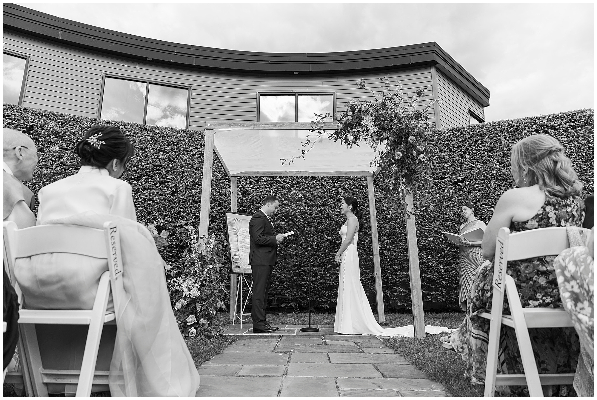 A bride and groom stand under a floral arch, exchanging vows outdoors at their roundhouse wedding. Guests are seated along a stone path in front of a modern building and tall hedge.