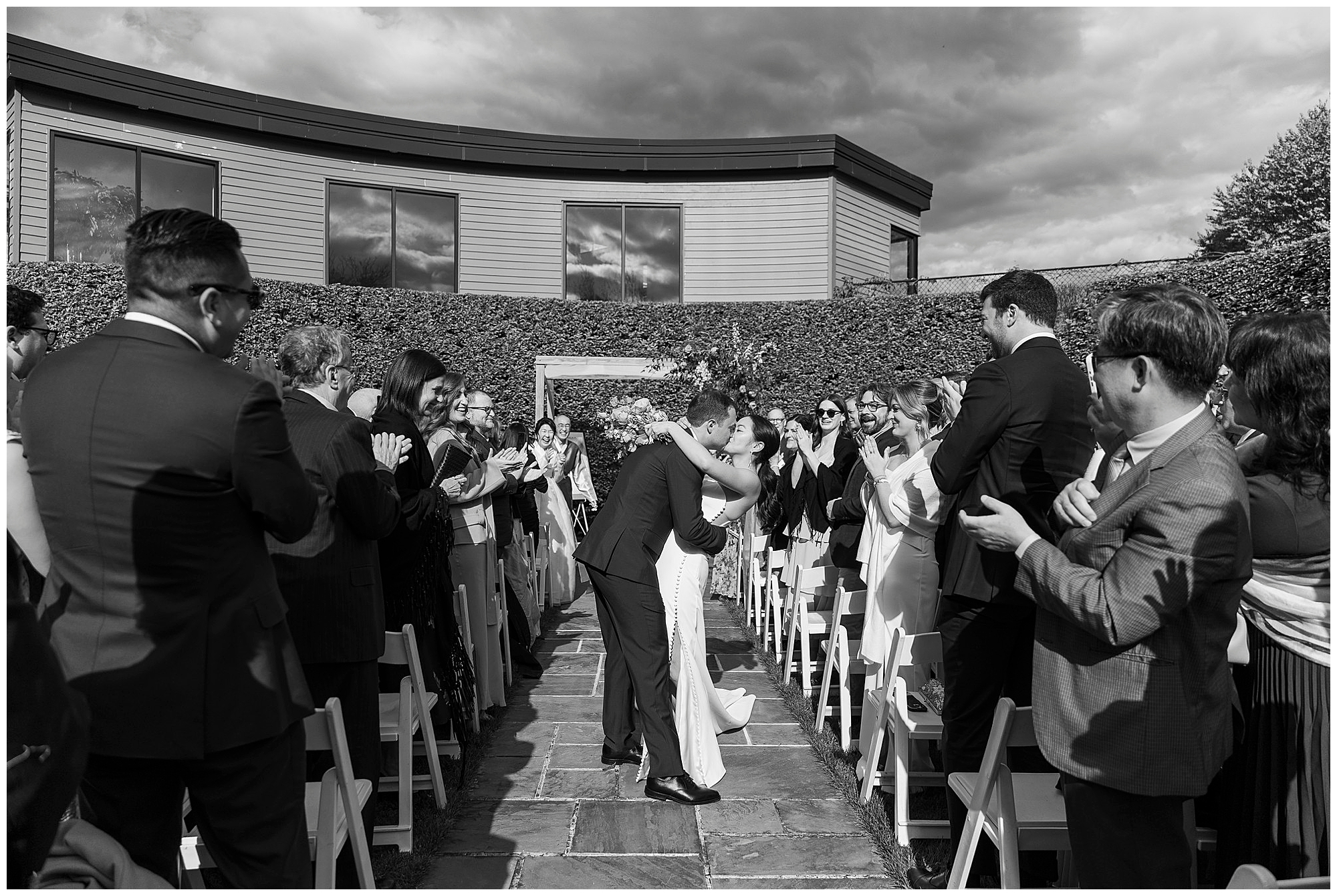 A newlywed couple kisses at the end of an outdoor aisle during their roundhouse wedding, surrounded by clapping guests on both sides under the sunny sky.