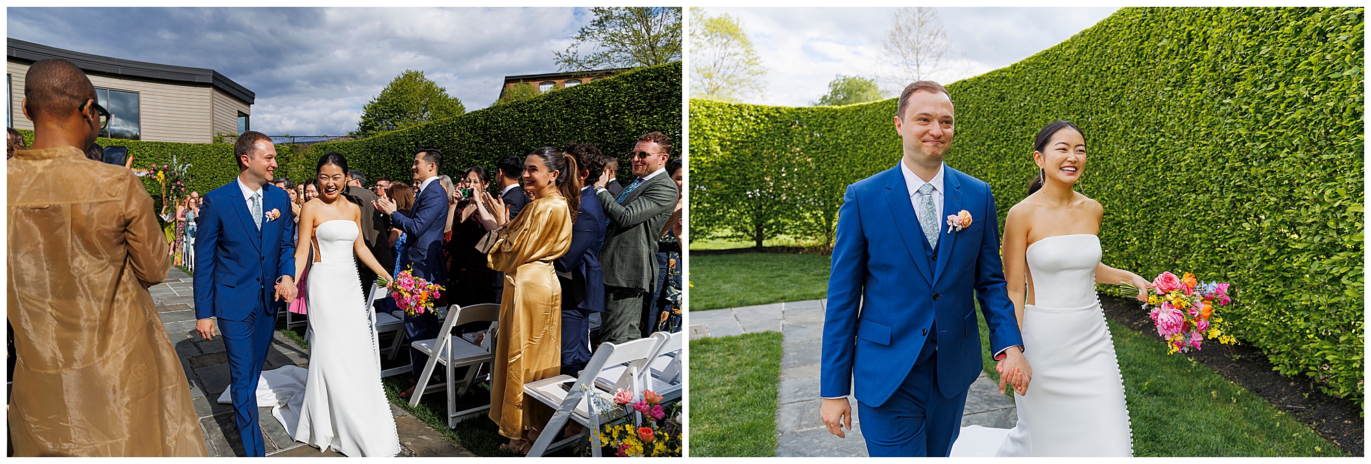 A bride in a white strapless dress and a groom in a blue suit walk down the outdoor aisle at their roundhouse wedding, smiling and holding hands, as guests take photos and celebrate beneath lush green hedges and a bright blue sky.