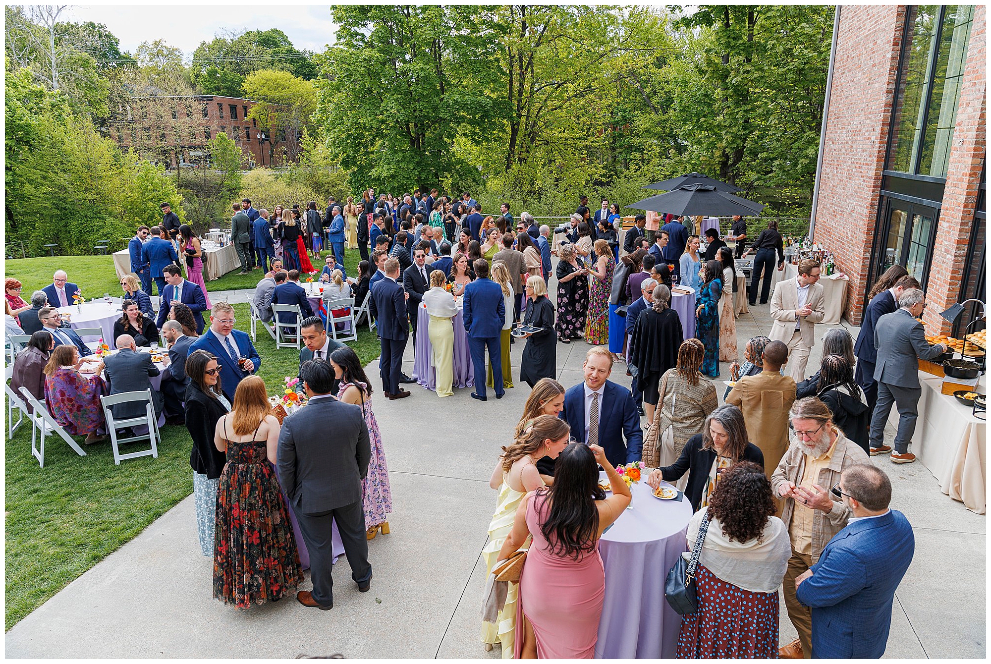 A large group of people in formal attire gather outdoors at a roundhouse wedding reception, standing and mingling around cocktail tables near a brick building with green trees in the background.