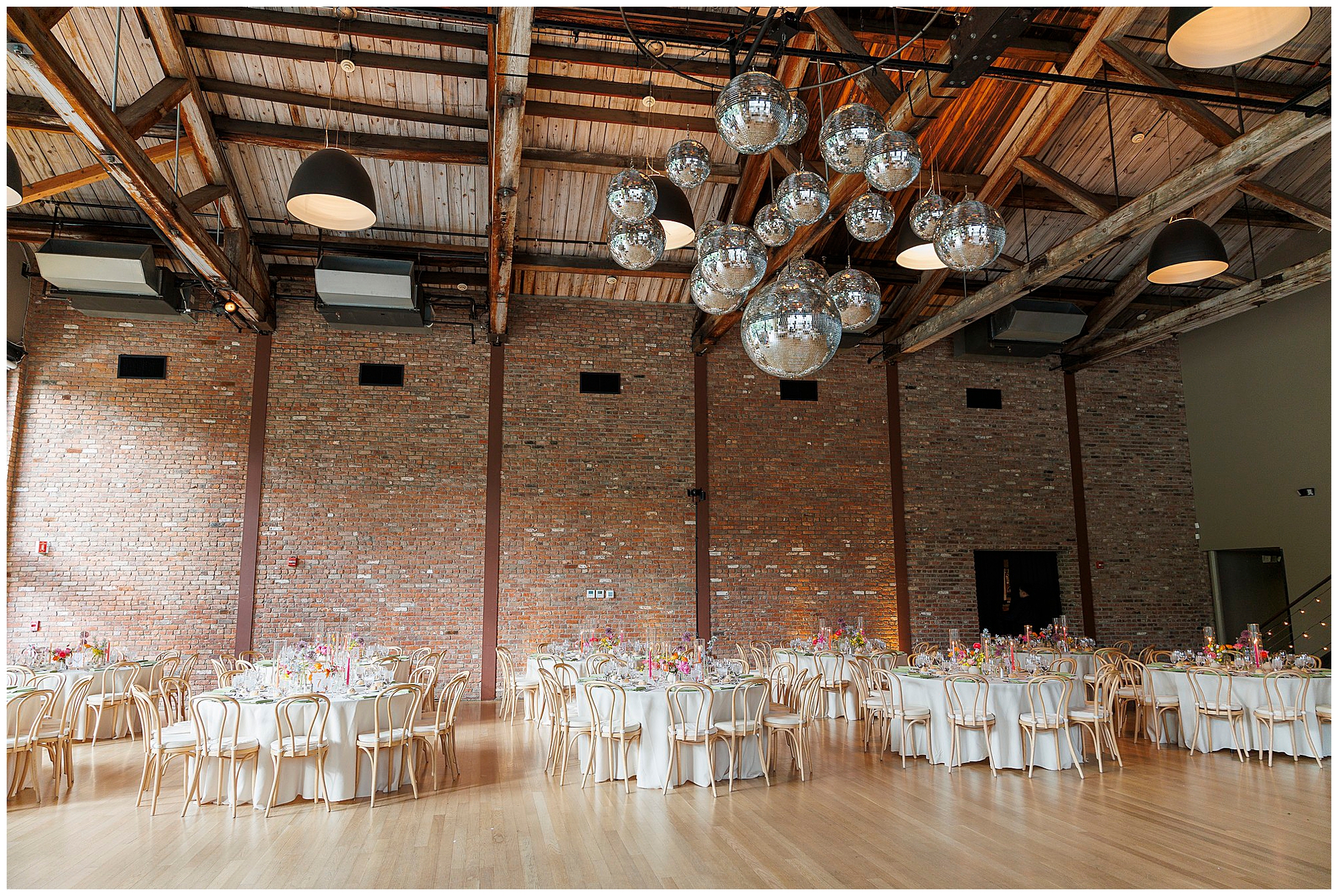A spacious event hall with exposed wooden beams, brick walls, and large hanging disco balls—perfect for a roundhouse wedding. Several round tables with white tablecloths and floral centerpieces are arranged for a celebration.