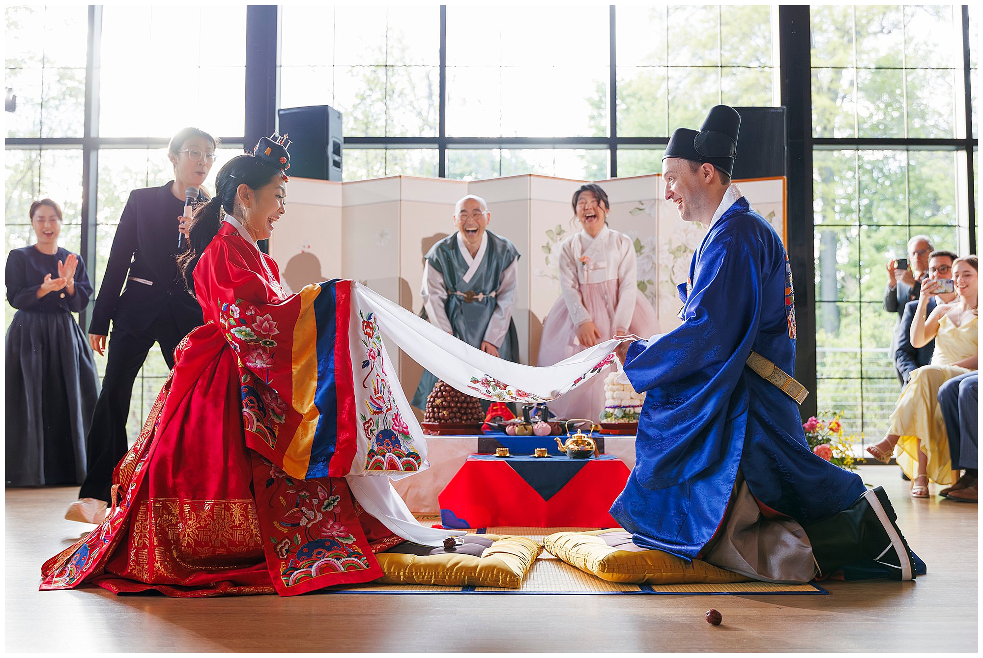 A bride and groom in traditional Korean hanbok kneel facing each other, holding a white cloth during a roundhouse wedding ceremony, with guests smiling and clapping in the background.