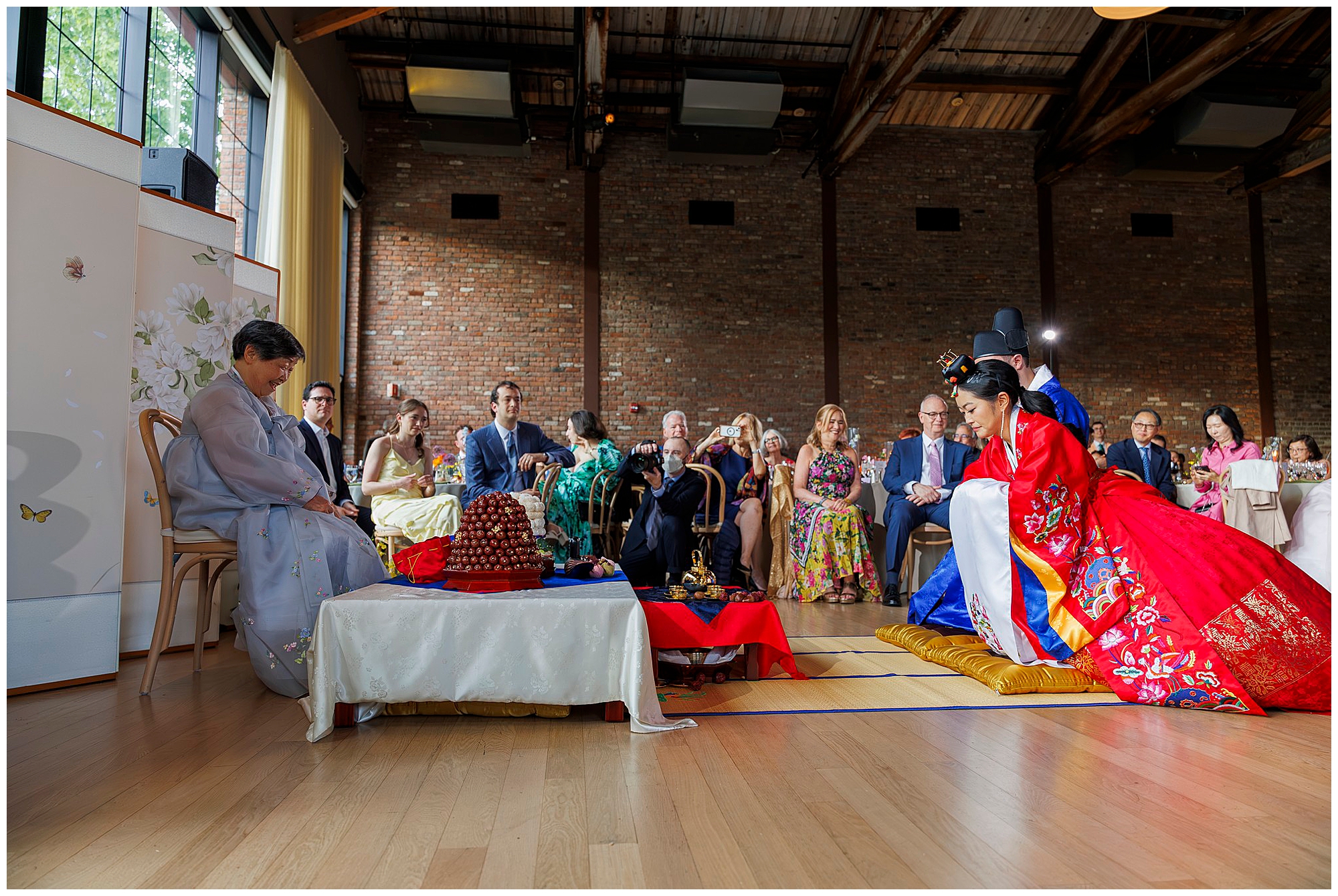 A couple in traditional Korean wedding attire sits facing each other on cushions during a ceremony at a roundhouse wedding, with guests seated and watching in a spacious hall with brick walls and large windows.