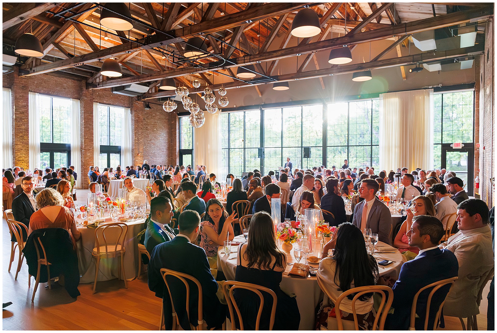 A large group of people sit at round tables decorated with flowers during a formal roundhouse wedding in a spacious, sunlit venue with high wooden ceilings and large windows.
