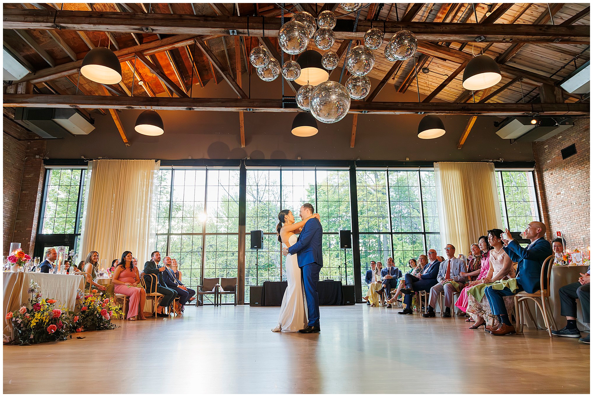 A bride and groom share their first dance in a sunlit, rustic roundhouse wedding venue with high wooden ceilings and large windows, while wedding guests watch from their seats surrounding the dance floor.