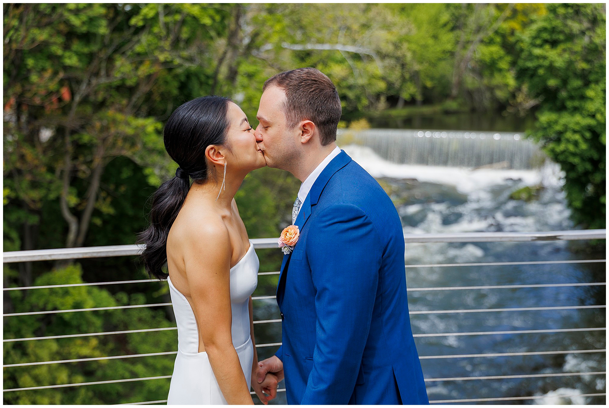 A bride and groom share a kiss on a bridge at their roundhouse wedding, surrounded by a flowing river and lush green trees. The bride wears a white strapless dress while the groom dons a royal blue suit with a boutonnière.
