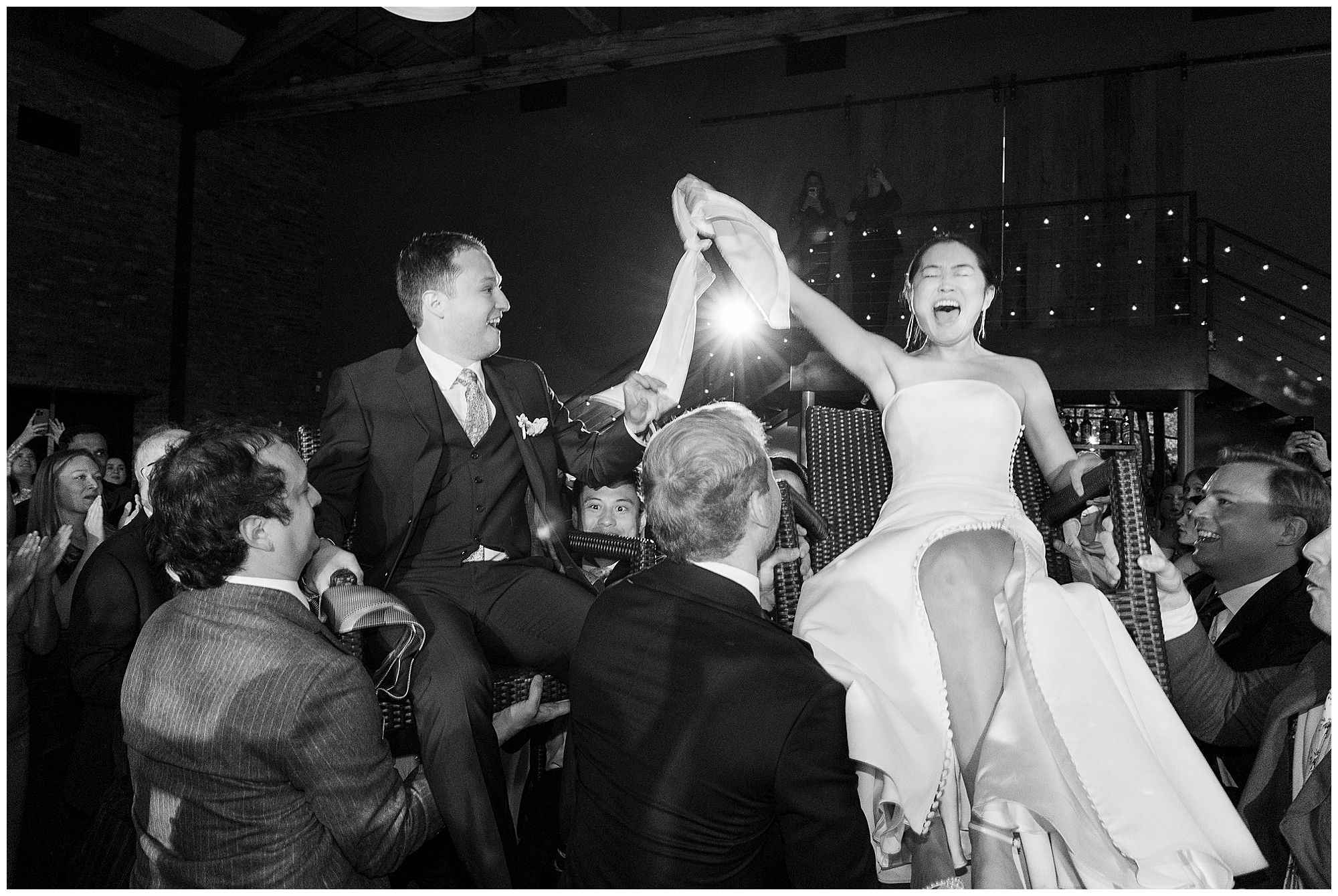 A bride and groom are joyfully lifted on chairs by guests during a roundhouse wedding celebration, holding hands and smiling. The bride laughs exuberantly as people cheer around them in a festive atmosphere.