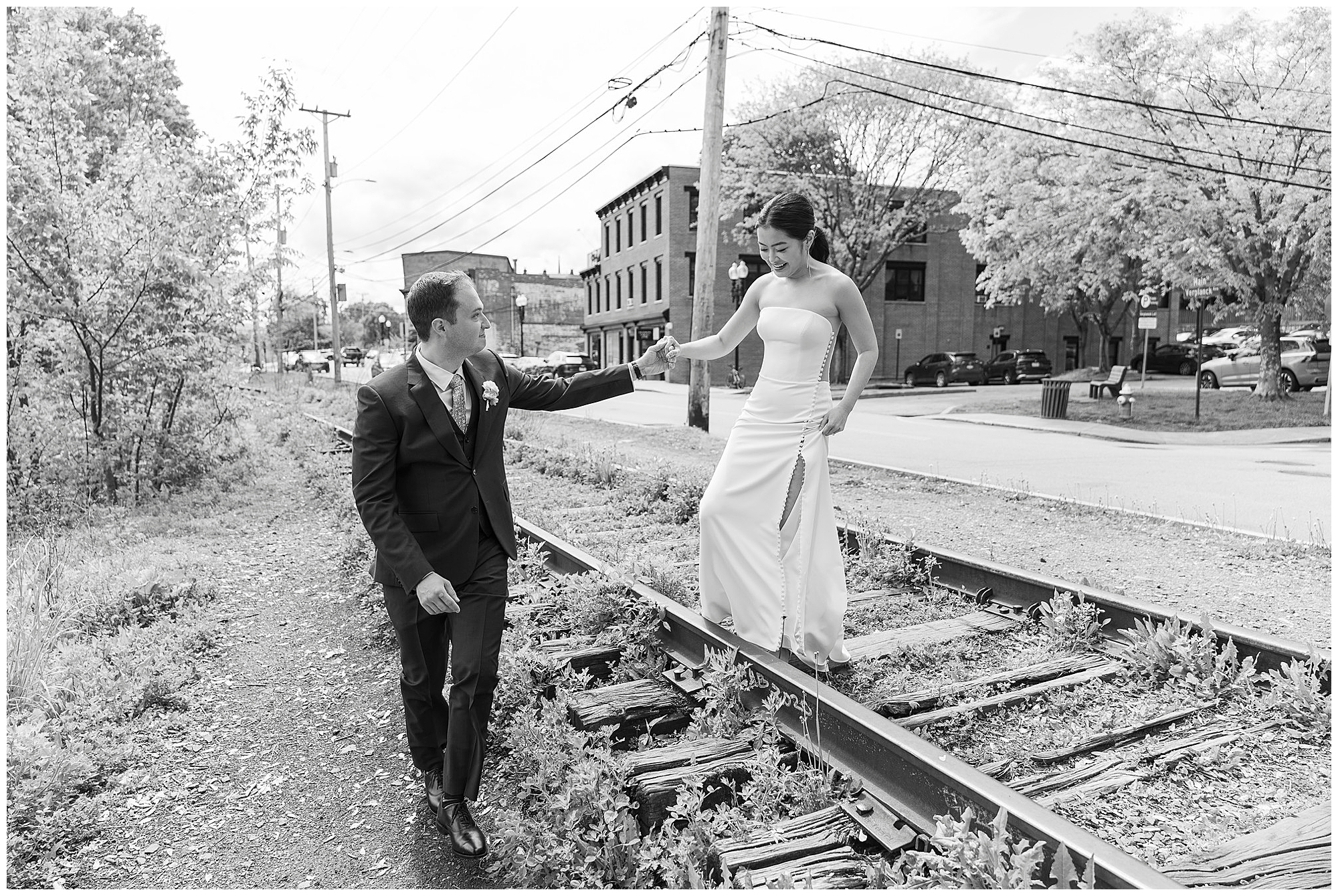 A groom in a suit holds the hand of a bride in a strapless dress as she walks along railroad tracks near an urban roundhouse wedding, surrounded by trees and city buildings.