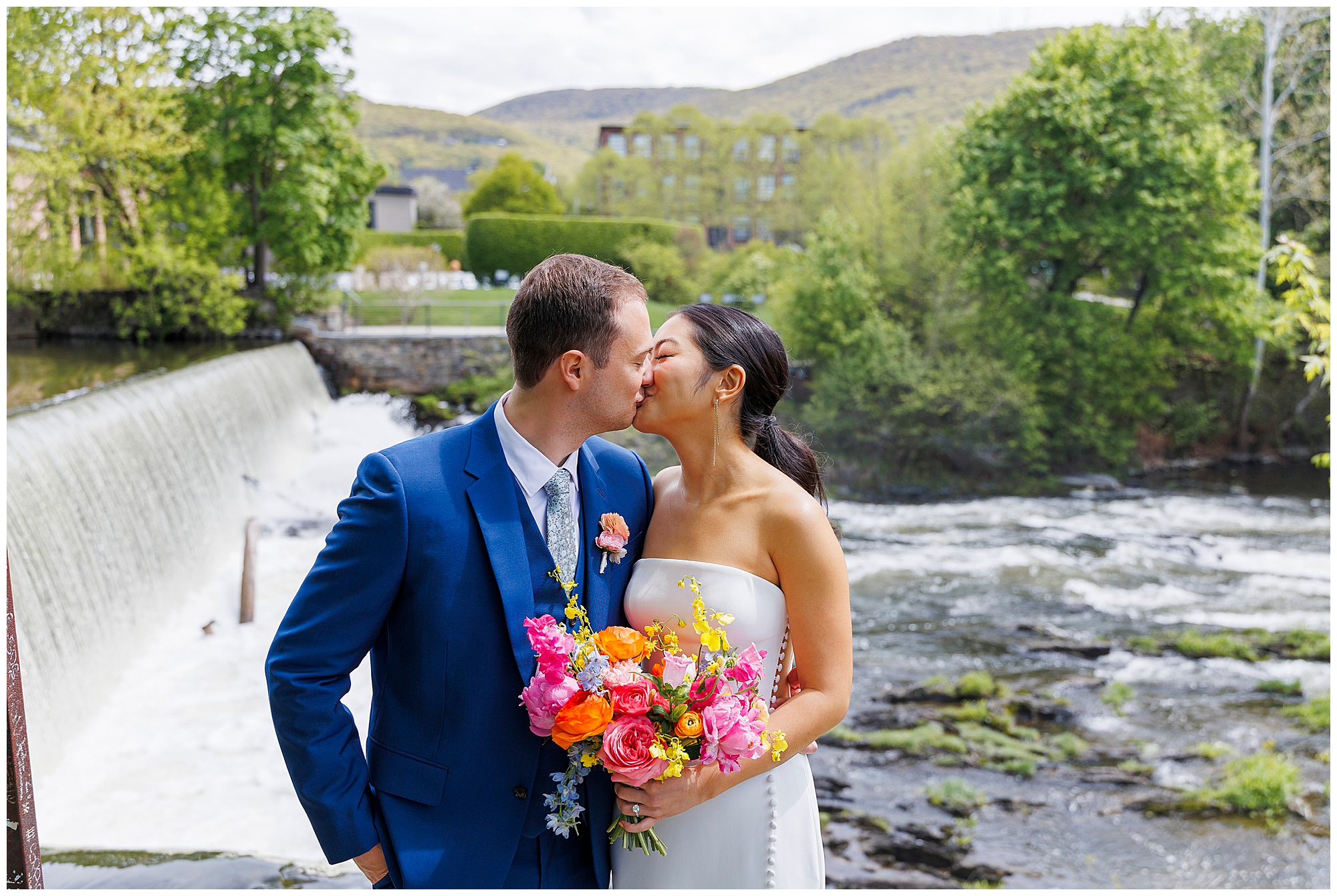 A couple in wedding attire kisses beside a river and small waterfall after their roundhouse wedding. The groom wears a blue suit and holds colorful flowers; the bride wears a strapless white dress, with green trees and distant hills in the background.