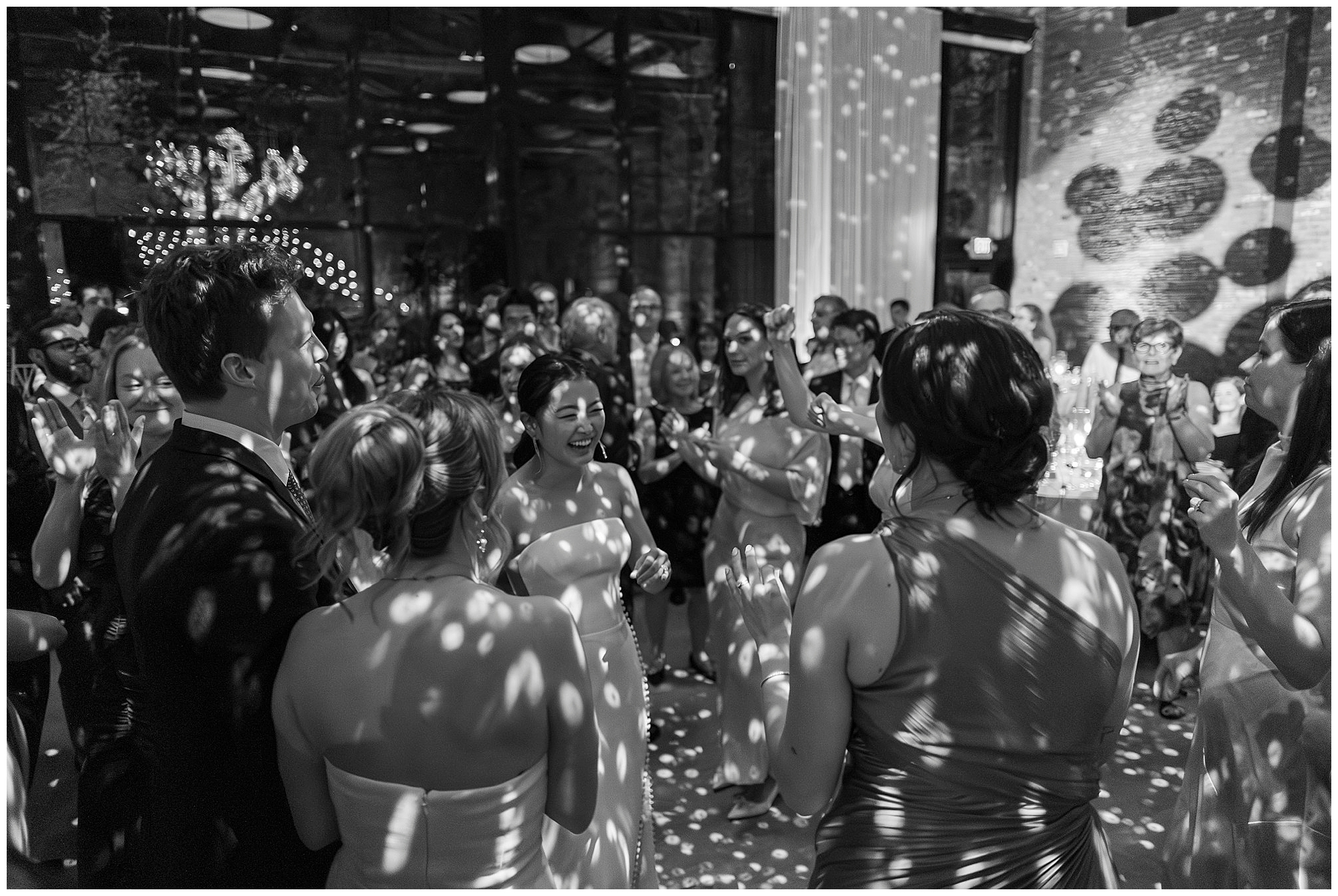 A joyful bride laughs surrounded by guests and bridesmaids on a lively, spotlit dance floor at a roundhouse wedding reception. Everyone appears to be celebrating together in an elegant venue.