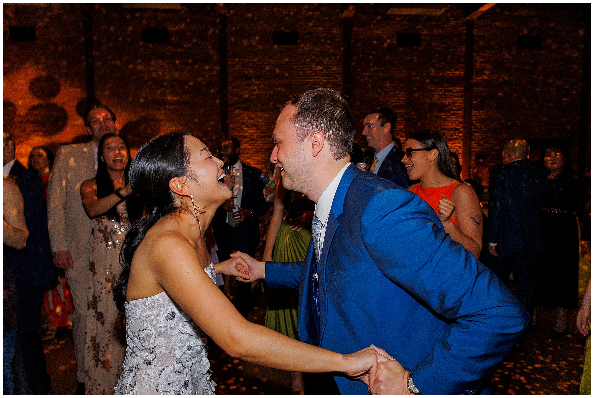 A couple joyfully dances together at a lively roundhouse wedding, surrounded by smiling guests in formal attire, with warm lighting and a brick wall in the background.