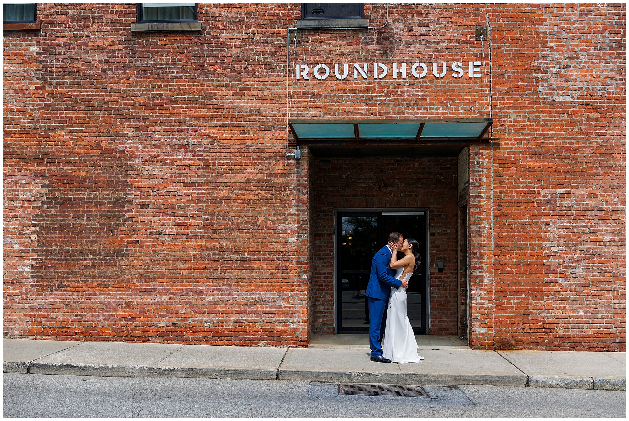 A couple dressed in wedding attire embraces and kisses under a glass awning at the entrance of the Roundhouse, capturing a romantic roundhouse wedding moment.