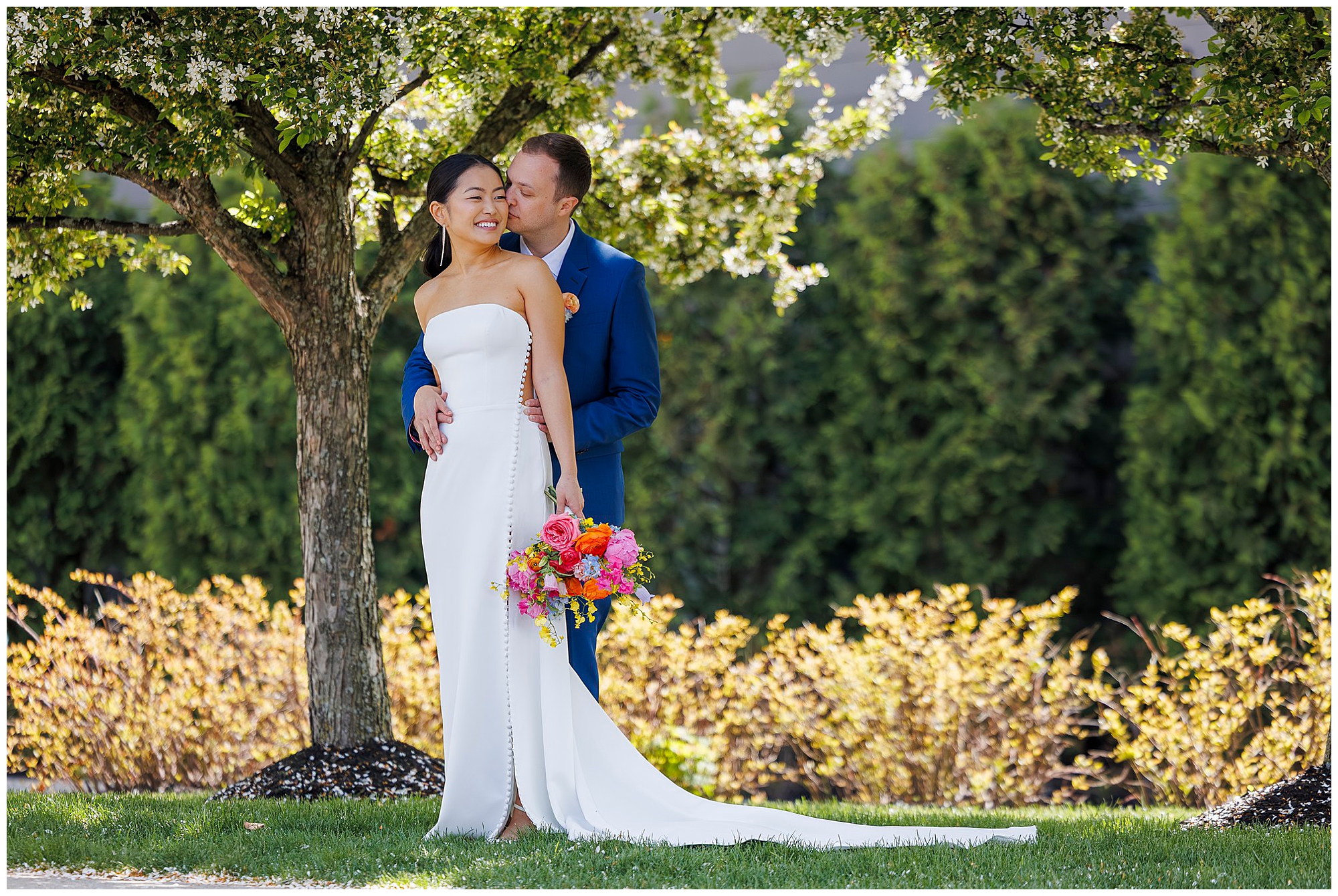 A bride in a white dress holds a colorful bouquet and smiles as the groom in a blue suit kisses her temple during their roundhouse wedding, standing under a blossoming tree with green foliage in the background.