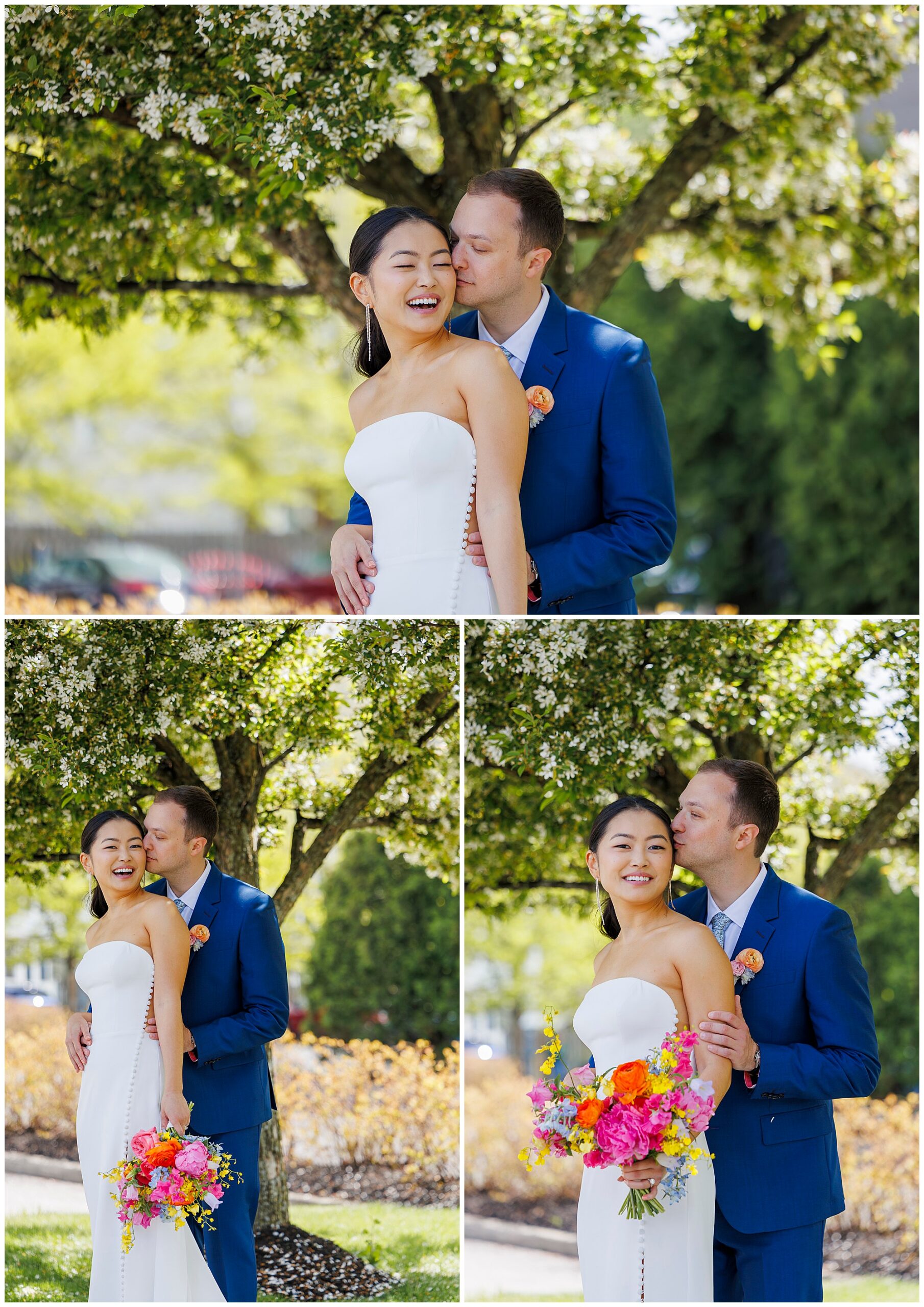A bride in a strapless white gown and a groom in a blue suit smile and embrace under a blooming tree at their roundhouse wedding. The bride holds a bright bouquet of flowers as both appear joyful and happy outdoors.