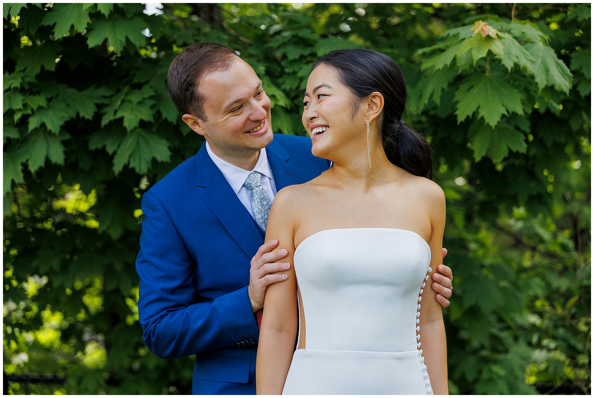 A smiling couple stands together outdoors at their roundhouse wedding; the man in a blue suit gently holds the woman in a strapless white dress as they look at each other against a backdrop of green leafy trees.