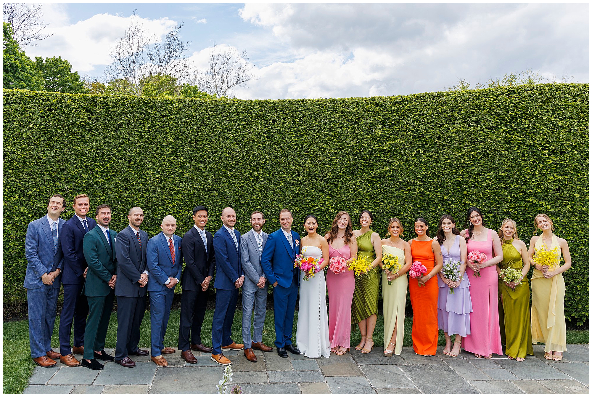A wedding party poses outdoors in front of a tall green hedge at a roundhouse wedding. The group includes the bride, groom, bridesmaids in colorful dresses, and groomsmen in suits, all smiling on a paved stone path under a partly cloudy sky.