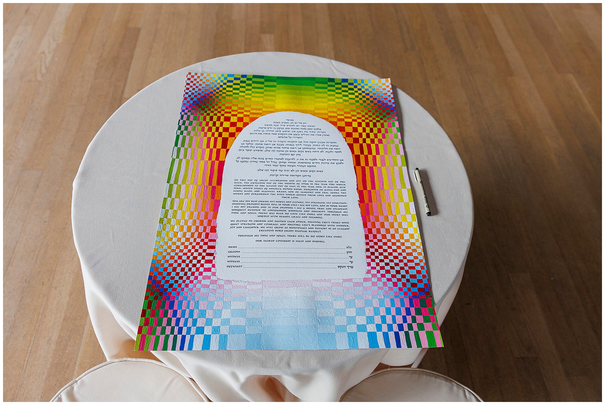A colorful, patterned document with Hebrew and English text lies on a round, white tablecloth-covered table on a wooden floor, evoking the charm of a roundhouse wedding. The paper features a vibrant rainbow checkerboard border.