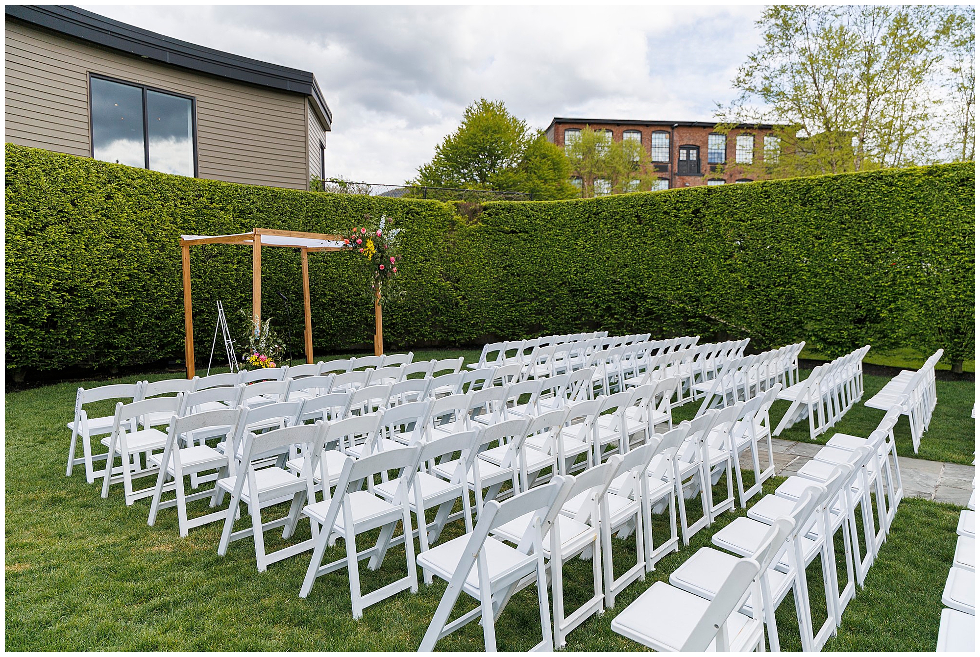 Rows of white folding chairs are arranged on a lawn, facing a simple wooden wedding arch adorned with flowers—perfect for a charming roundhouse wedding in a private garden framed by tall green hedges and nearby buildings.