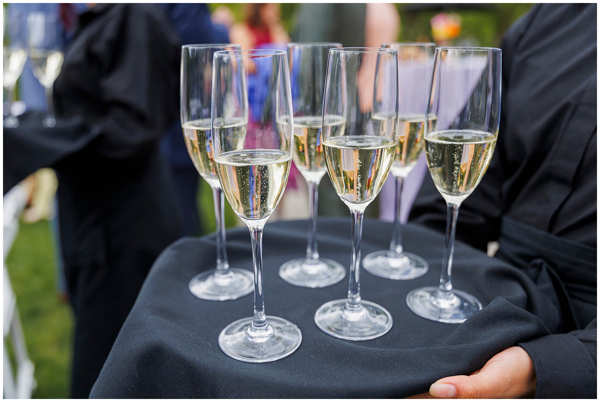 A server holding a tray with six filled champagne glasses, covered with a black cloth, at a roundhouse wedding. People are blurred in the background, suggesting a festive outdoor event.
