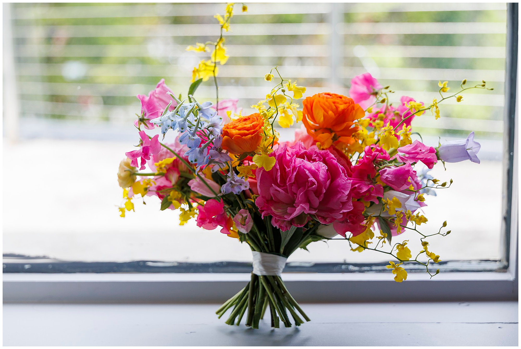 A colorful bouquet of pink, orange, yellow, and purple flowers sits on a windowsill with a blurred outdoor background—perfect for a roundhouse wedding. The stems are tied together with white ribbon.