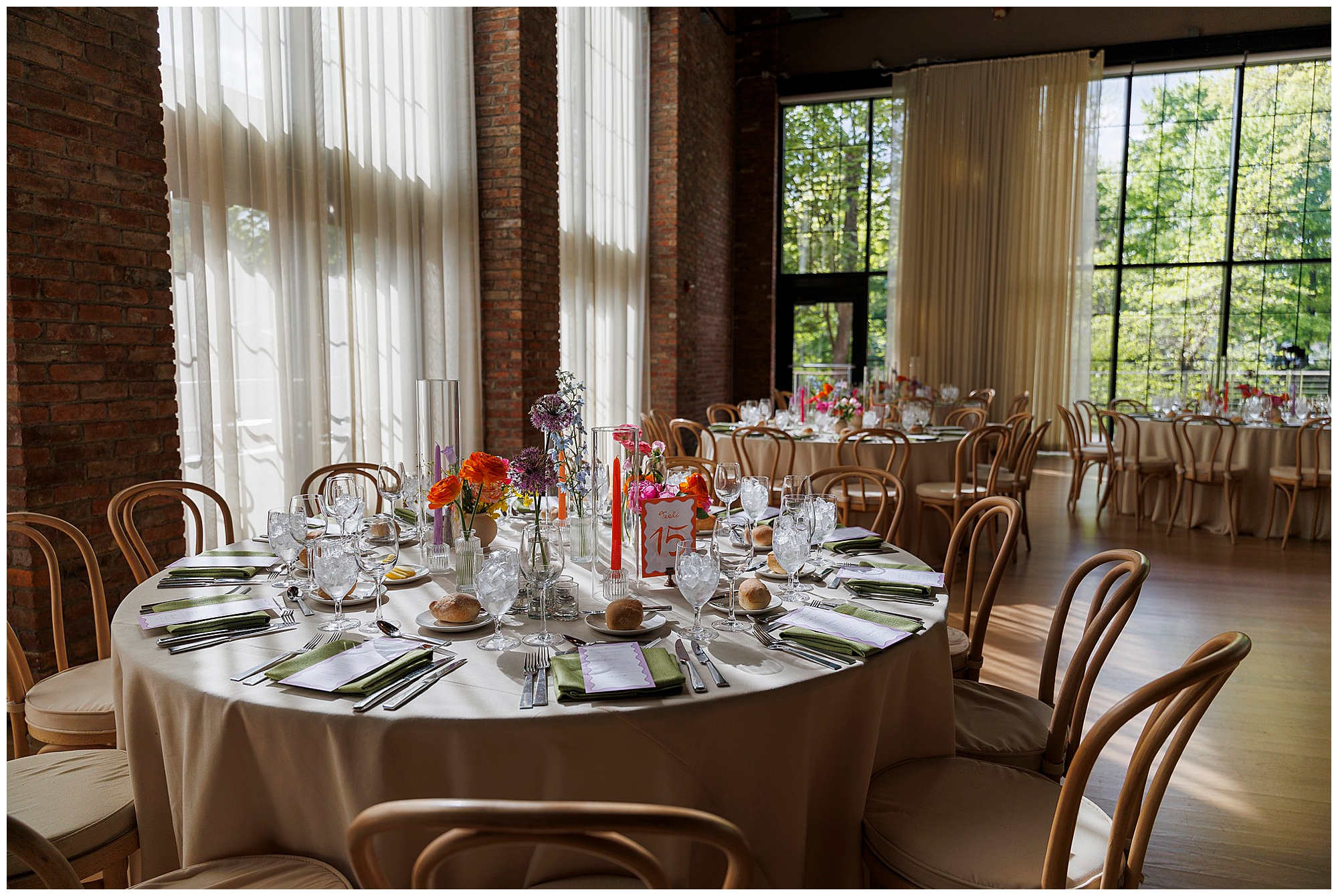 Elegant round table set for a formal event with floral centerpieces, glassware, and napkins, surrounded by wooden chairs in a sunlit room with large windows and brick walls—the perfect setting for a sophisticated roundhouse wedding.