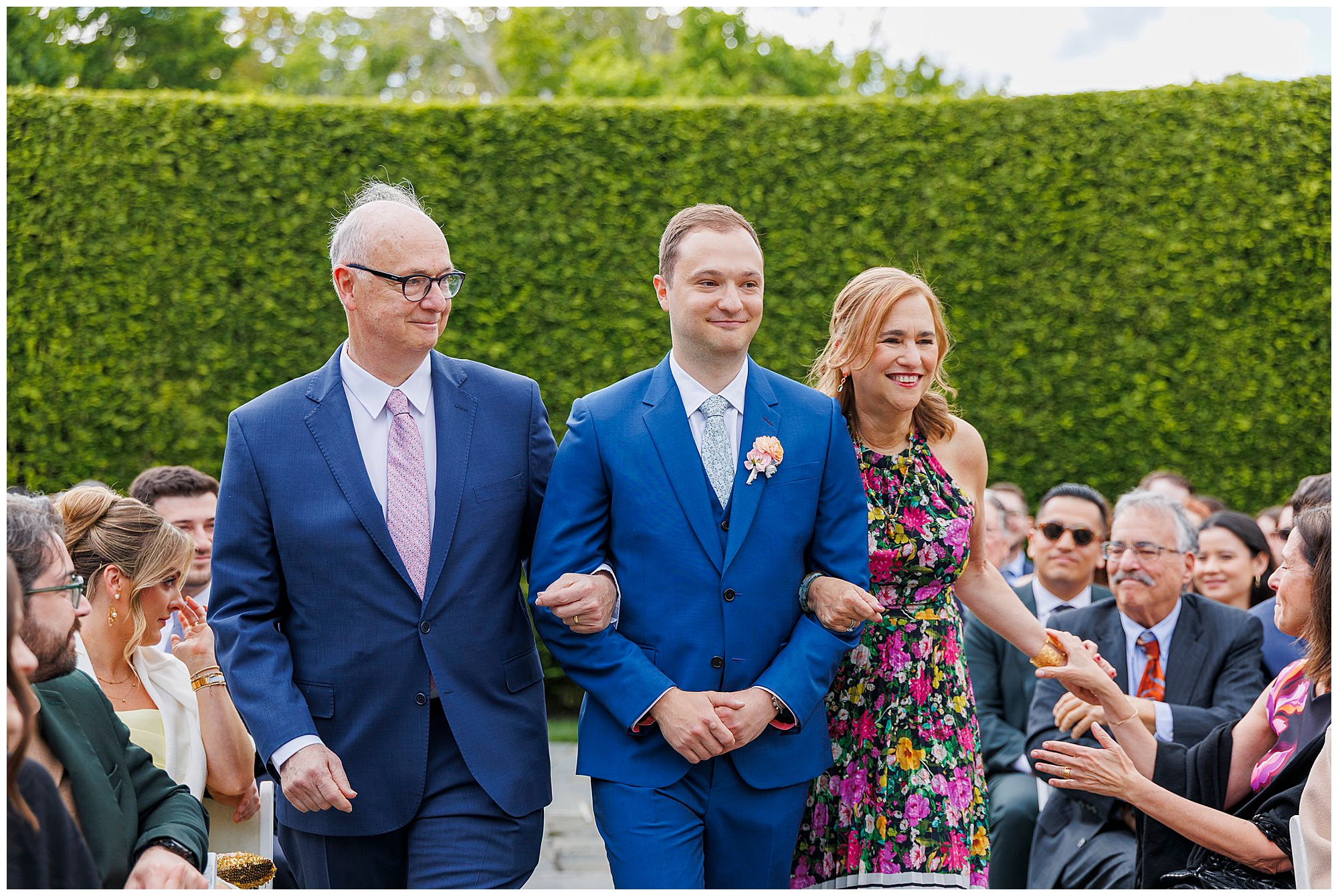 A smiling man in a blue suit walks between an older man in a suit and a woman in a floral dress, holding their arms, as people seated on either side watch and smile outdoors at a roundhouse wedding.
