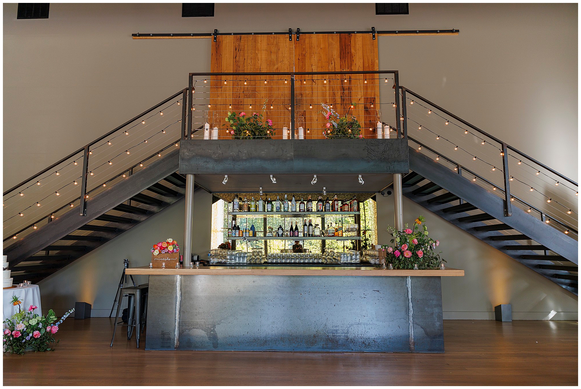 A modern indoor bar with a metal counter, shelves stocked with bottles, and floral arrangements. Perfect for a wedding at the Roundhouse, string-lit staircases flank the bar and lead up to a balcony with wooden doors and more flowers.