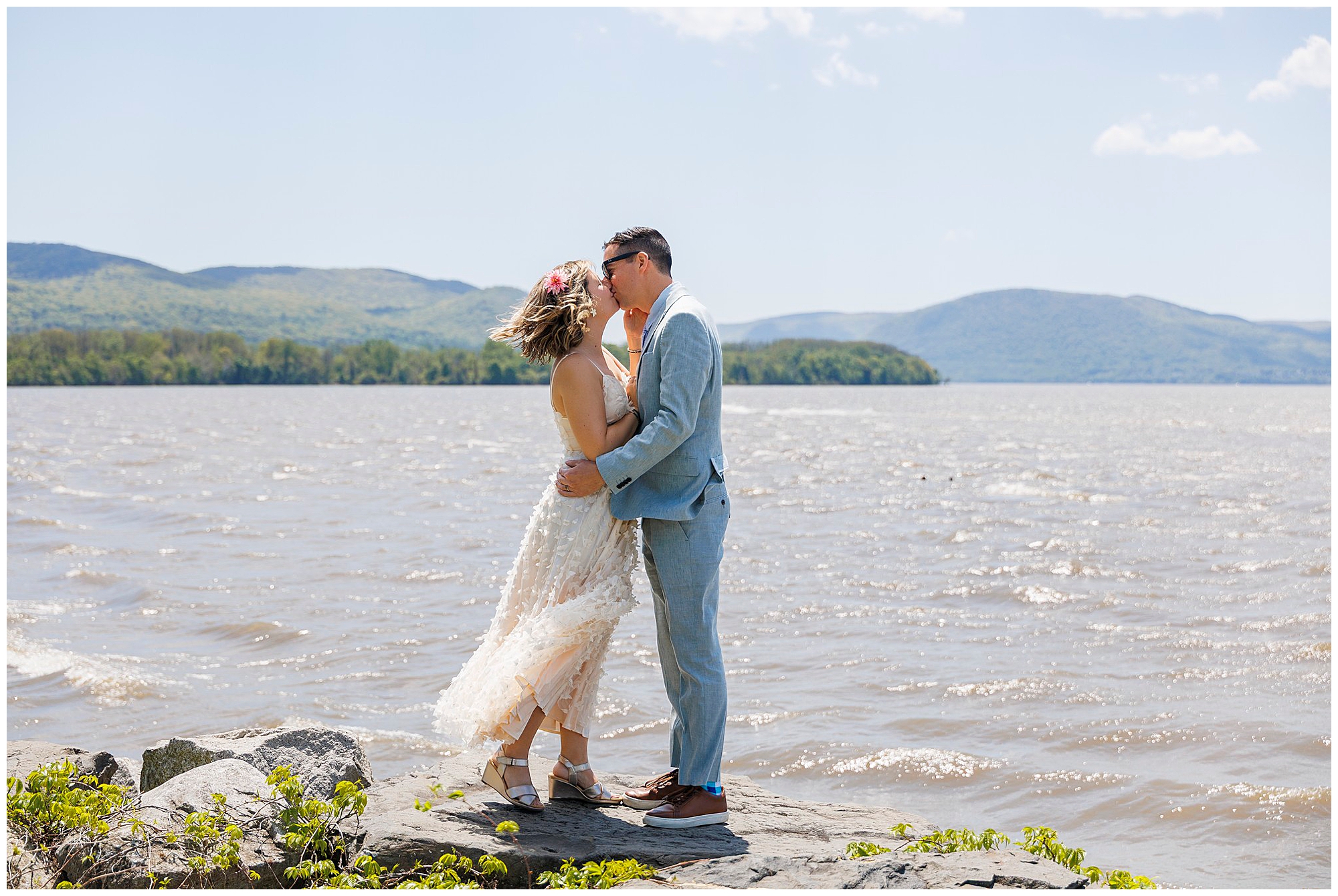 A couple in wedding attire stands on rocks by a lake, kissing after their Beacon Courthouse elopement. The bride wears a white dress and flower crown, the groom is in a light blue suit. Mountains and blue sky frame their special moment.