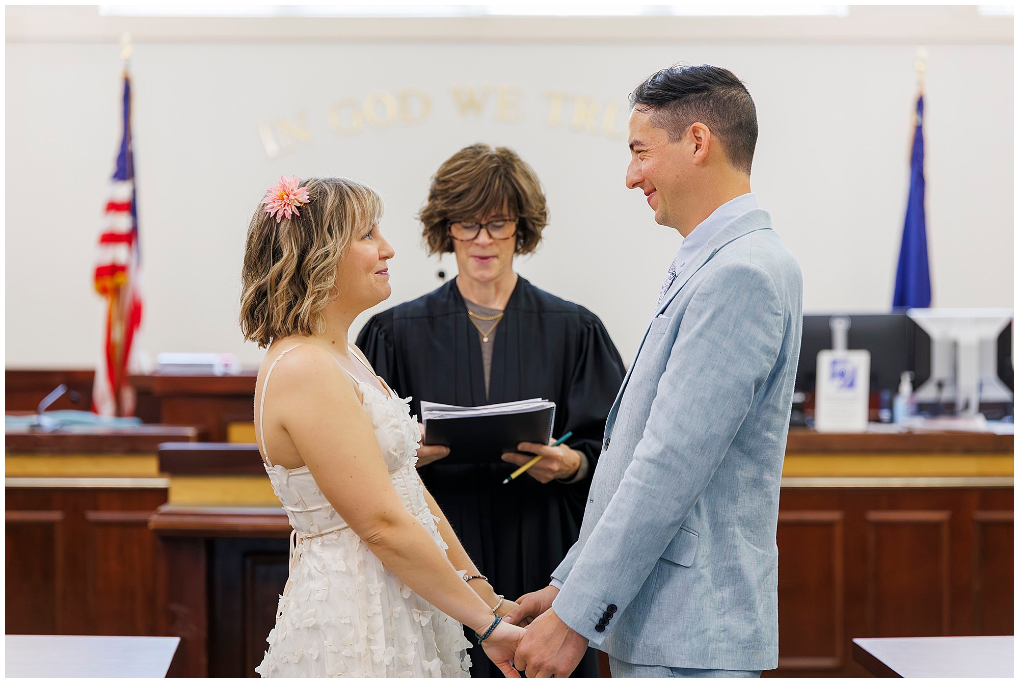 A couple stands holding hands, facing each other and smiling during a wedding ceremony in a courtroom, with a judge officiating behind them. American flags and a sign reading IN GOD WE TRUST are visible in the background.
