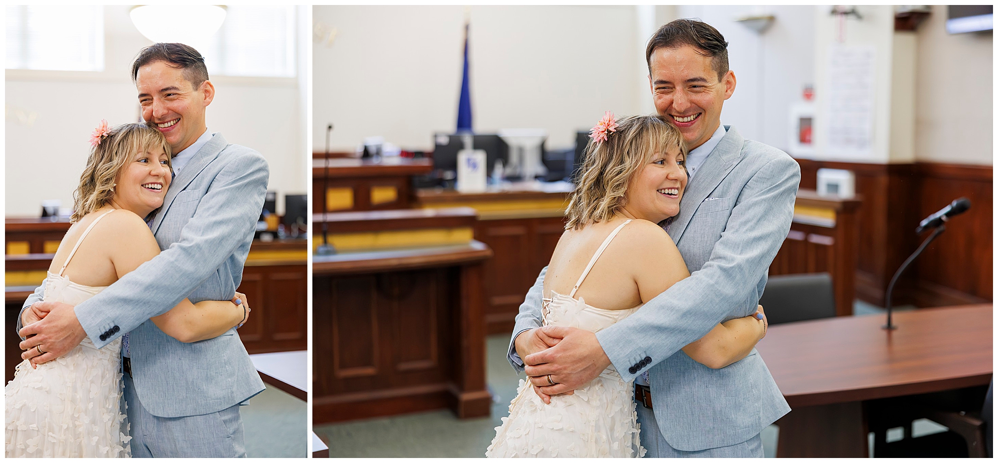 A joyful couple in wedding attire embraces and smiles in a courtroom, suggesting a courthouse wedding. The man wears a light blue suit, and the woman wears a white dress with pink flowers in her hair.