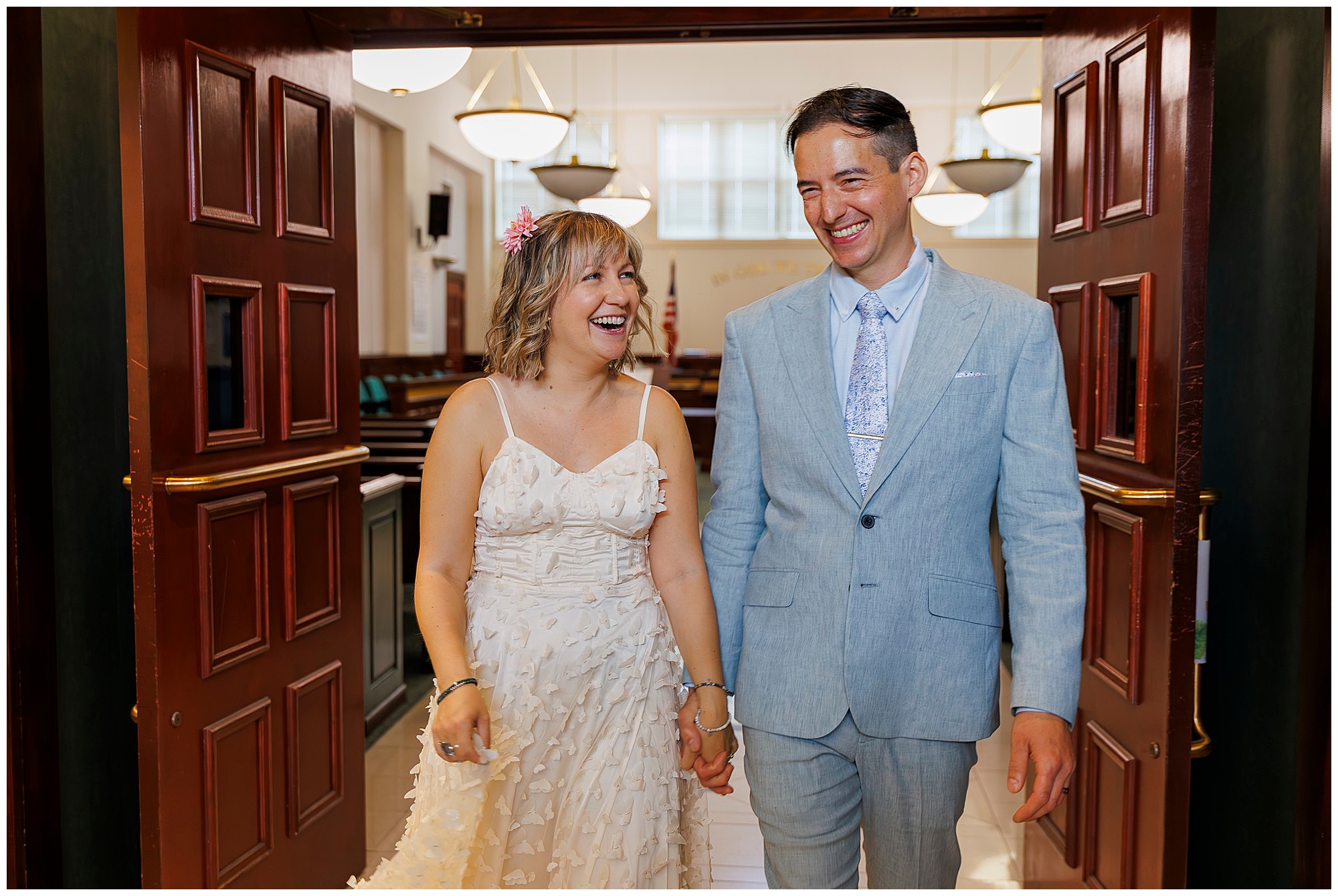 A joyful couple holding hands and smiling, walking through large wooden doors in a bright indoor setting. The woman wears a white dress with floral details, and the man is in a light blue suit and tie.