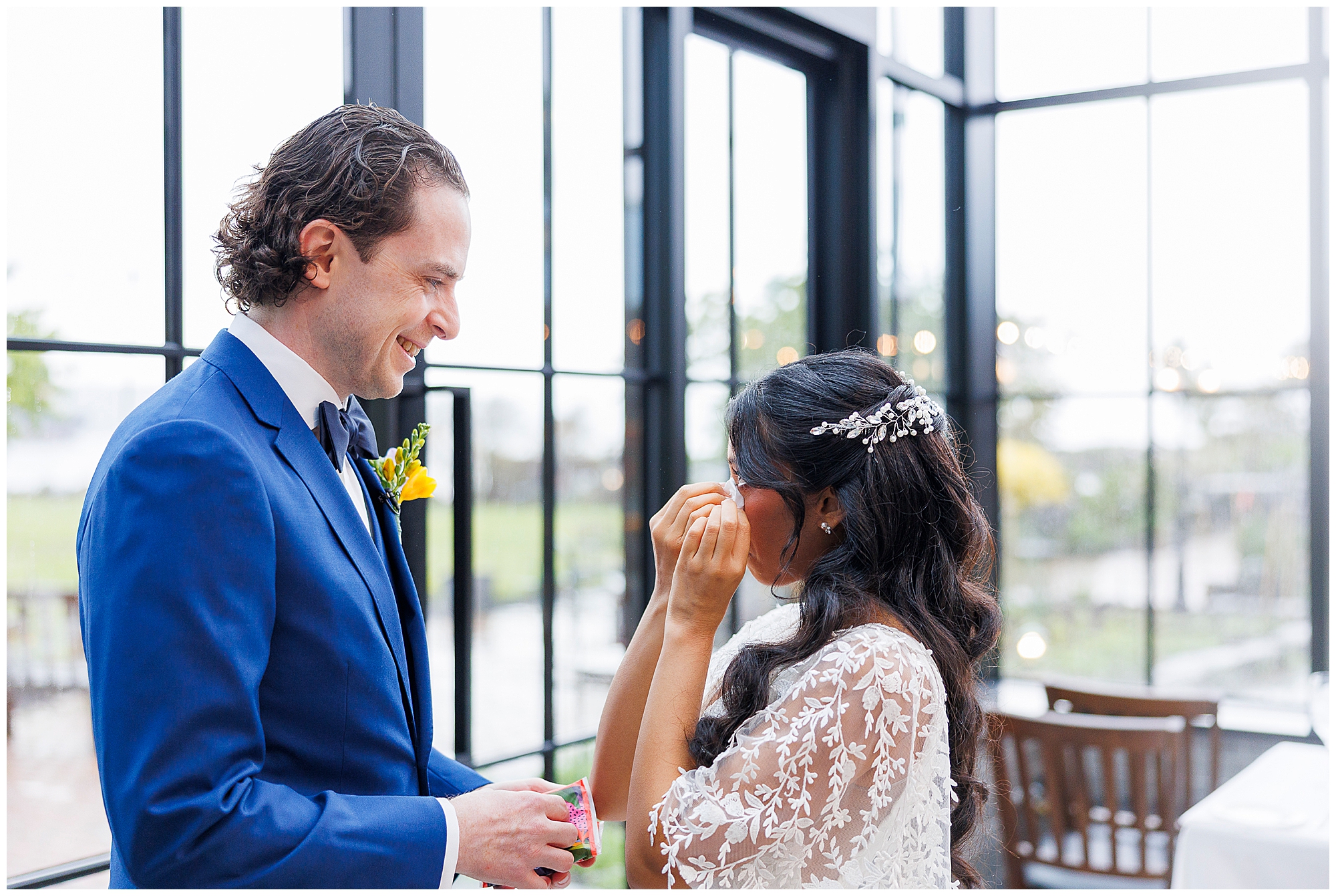 A groom in a blue suit smiles warmly at his bride, who is wiping away happy tears during their Harvest on Hudson micro wedding. She wears a lace dress and jeweled hairpiece as they stand together indoors by large windows framing lush greenery.