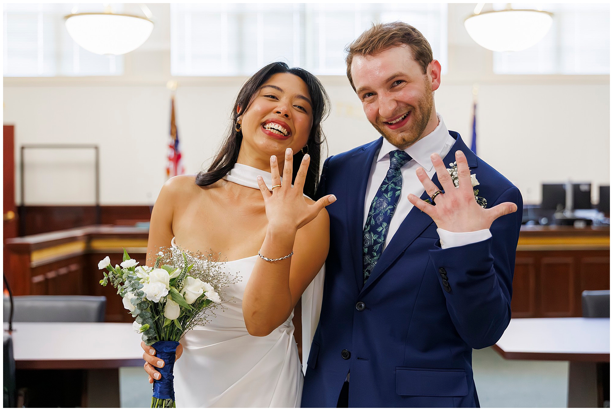 A joyful bride and groom in wedding attire show off their wedding rings, smiling at the camera. The bride holds white flowers as they stand in a bright, formal room with American flags—capturing their Beacon City Hall elopement.