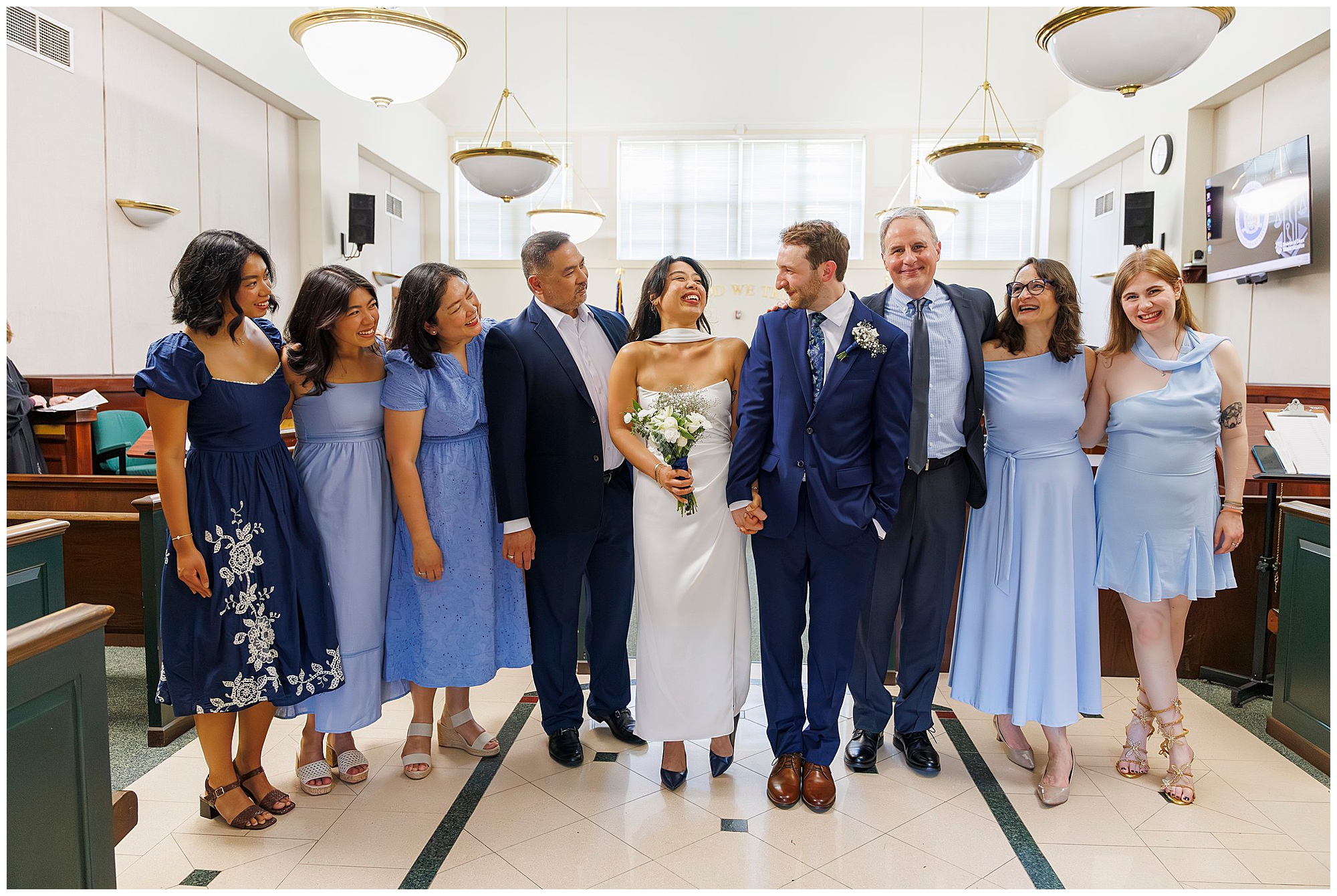 A bride and groom stand smiling in the center of family and friends, all dressed in blue and white attire, inside the brightly lit Beacon City Hall. Everyone appears happy and celebratory at this intimate elopement gathering.