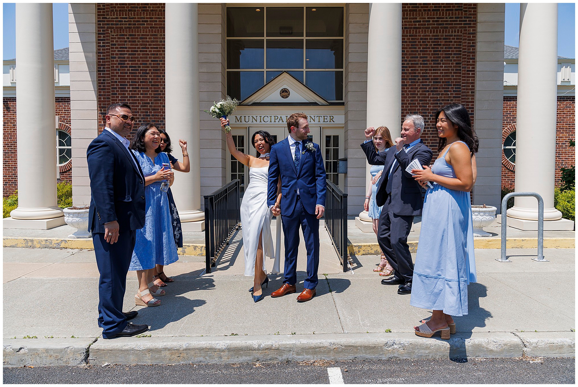 A bride and groom stand smiling outside Beacon City Hall after their elopement, surrounded by friends and family dressed in blue outfits, celebrating and cheering in the sunlight.