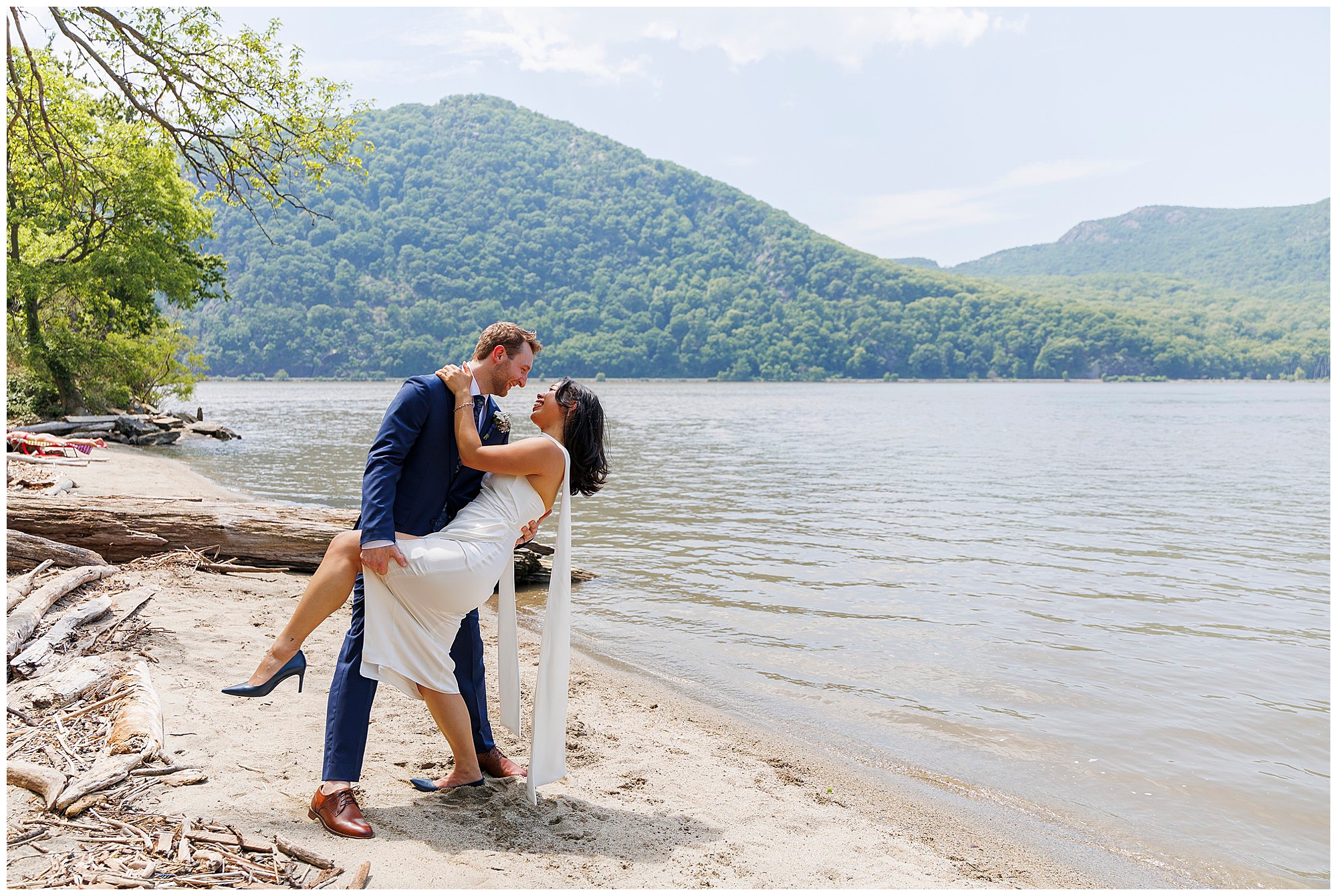 A couple dressed in wedding attire poses playfully on a sandy lakeshore, celebrating their Beacon City Hall elopement. The man dips the woman as green forested hills and calm water stretch behind them under a partly cloudy sky.