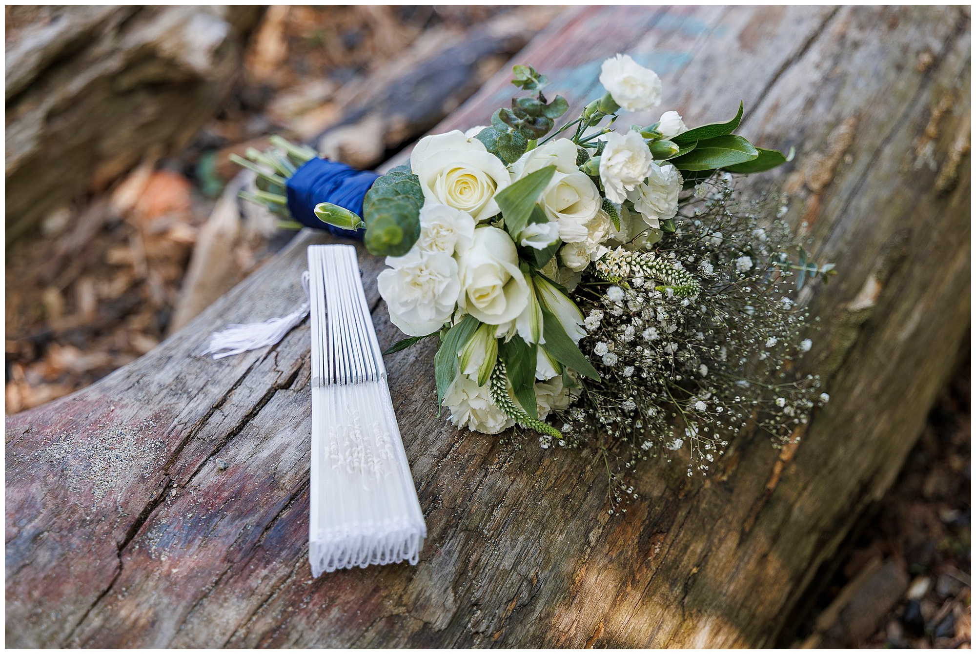 A bouquet of white roses and greenery rests on a weathered log outdoors, beside a bundle of folded white paper fans with tassels—a lovely touch for a Beacon City Hall elopement.