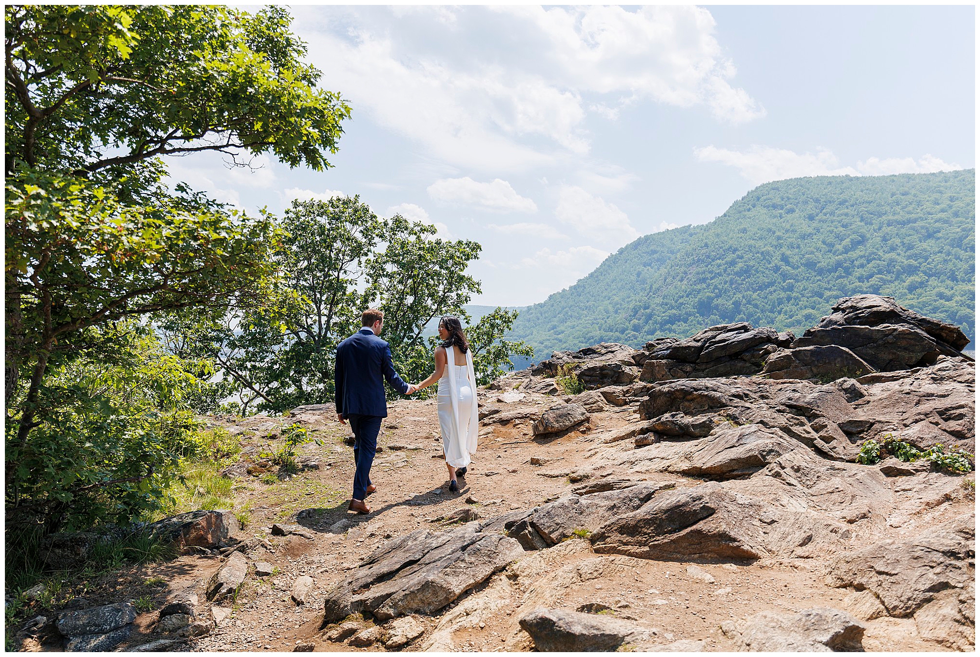A couple, dressed formally, walk hand in hand on a rocky trail surrounded by trees and mountains under a bright, partly cloudy sky after their intimate Beacon City Hall elopement.