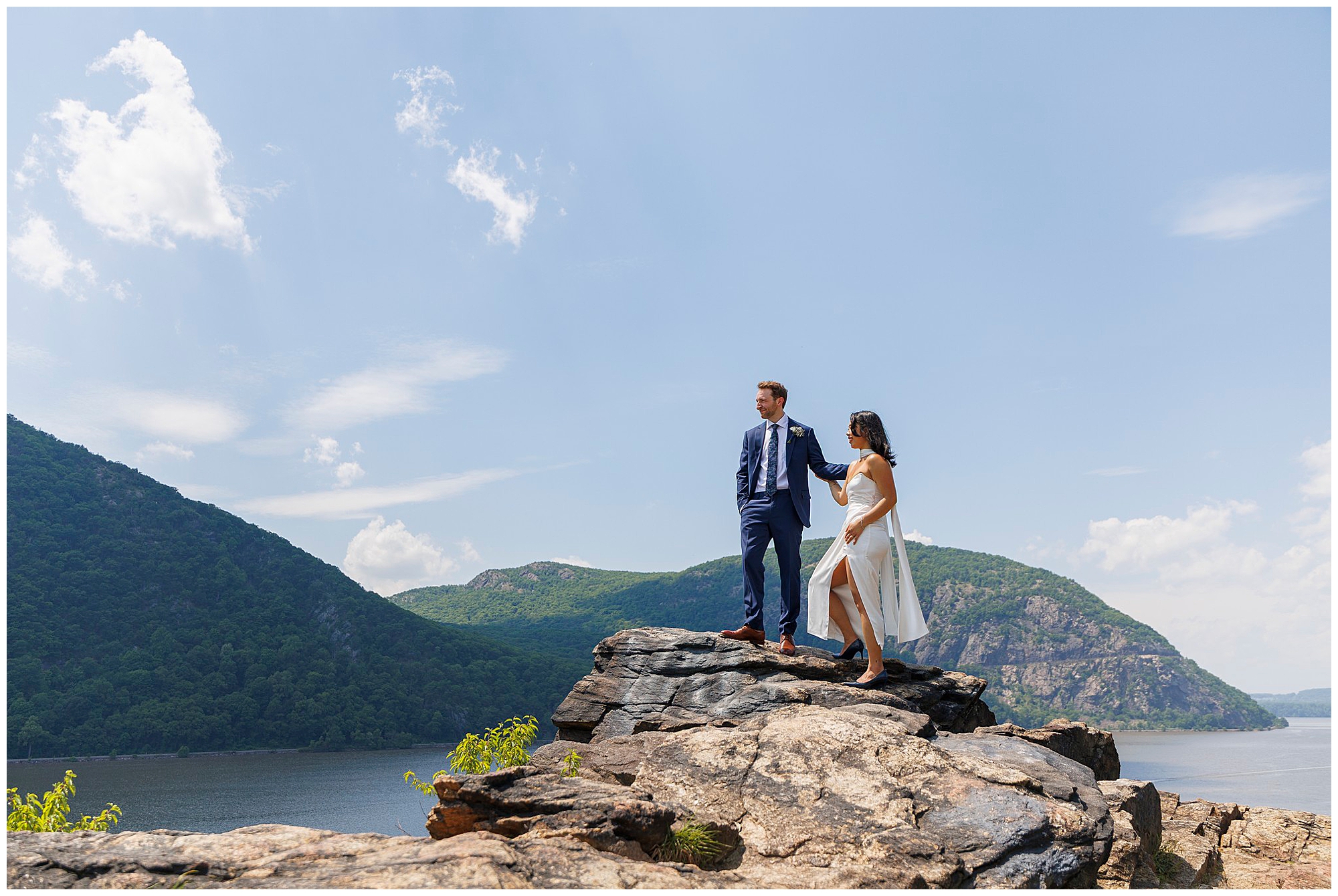 A couple stands on rocky cliffs overlooking a river and green mountains after their Beacon City Hall elopement. The man wears a suit and the woman a white dress under a clear sky, creating a scenic, adventurous atmosphere.
