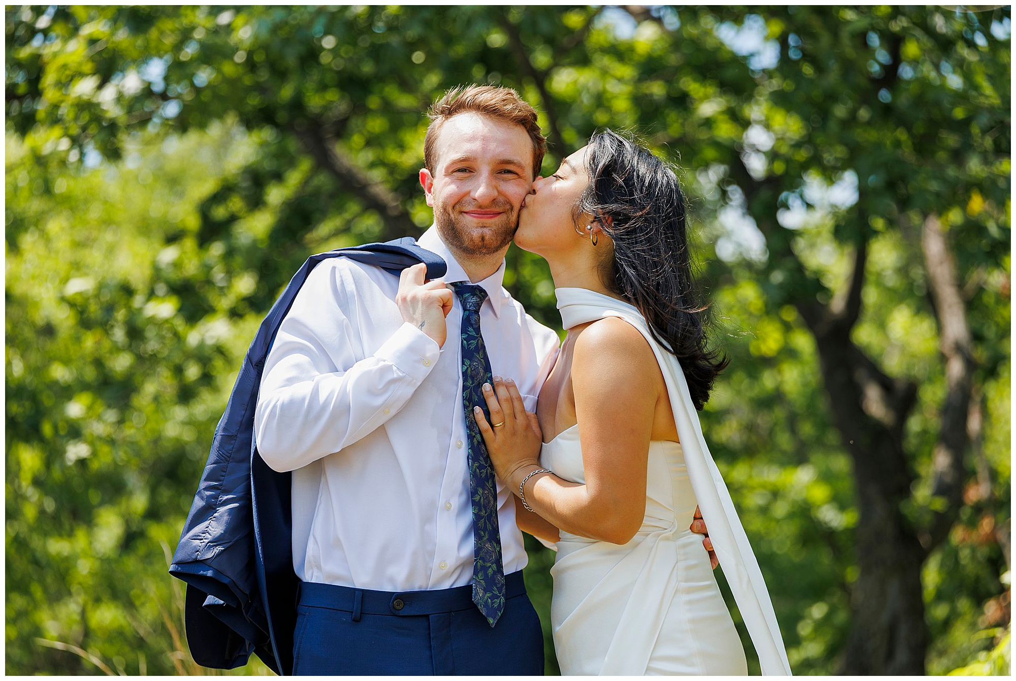 A woman in a white dress kisses a smiling man on the cheek outdoors after their Beacon City Hall elopement. The man holds a jacket over his shoulder and wears a white shirt and blue tie, with green trees filling the background.